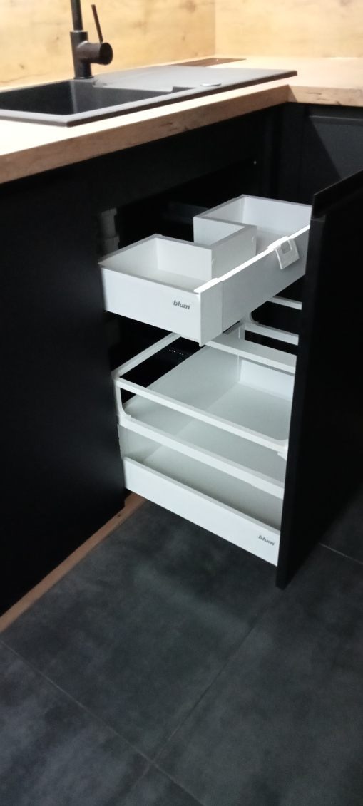 Wysuwane, białe szuflady z organizerami w czarnej zabudowie kuchennej, widoczne logo Blum, szare płytki podłogowe.