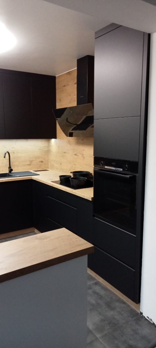 Nowoczesna kuchnia z czarnymi matowymi frontami, drewnianym blatem i okładziną ścienną, czarnym zlewem i armaturą, oraz czarnym piekarnikiem i okapem.