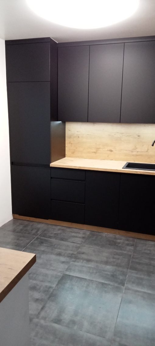 Ciemna zabudowa kuchenna z drewnianym blatem i czarnym zlewem, oświetlenie LED pod szafkami, szare płytki podłogowe.