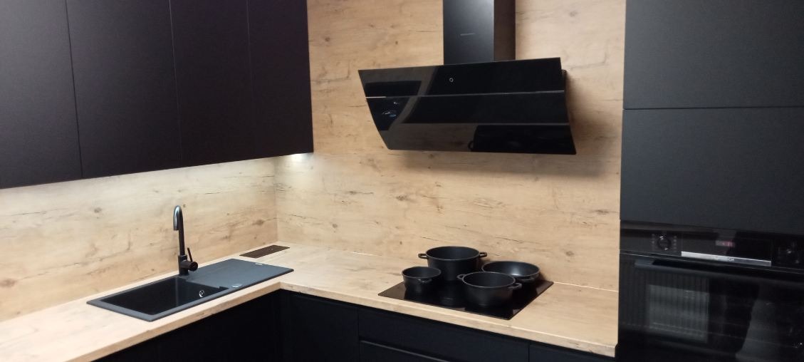 Część czarnej zabudowy kuchennej z drewnianym blatem, czarnym zlewozmywakiem, baterią i płytą indukcyjną z czarnymi garnkami, podświetlona listwą LED pod szafkami.