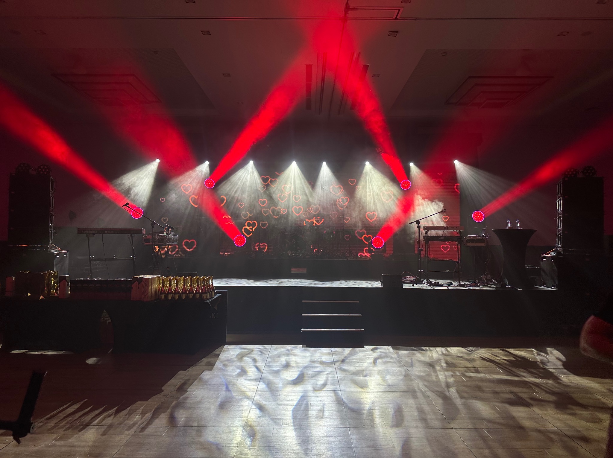 Scena oświetlona czerwonymi reflektorami z motywem serc, przygotowana na imprezę, z widocznymi instrumentami muzycznymi i nagrodami na stole.