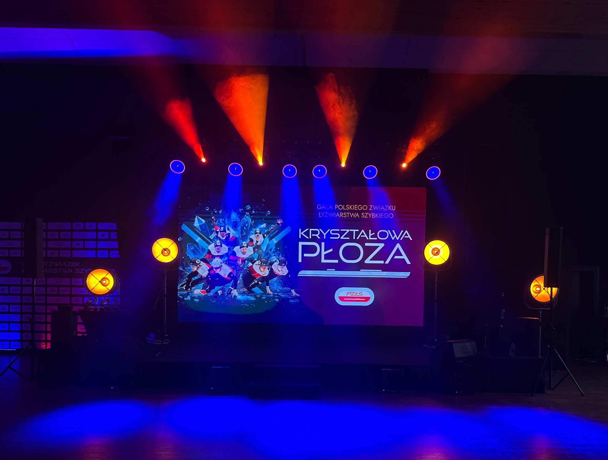 Scena z ekranem wyświetlającym 'Gala Polskiego Związku Łyżwiarstwa Szybkiego Kryształowa Płoza', oświetlona reflektorami w kolorach pomarańczowym i niebieskim.