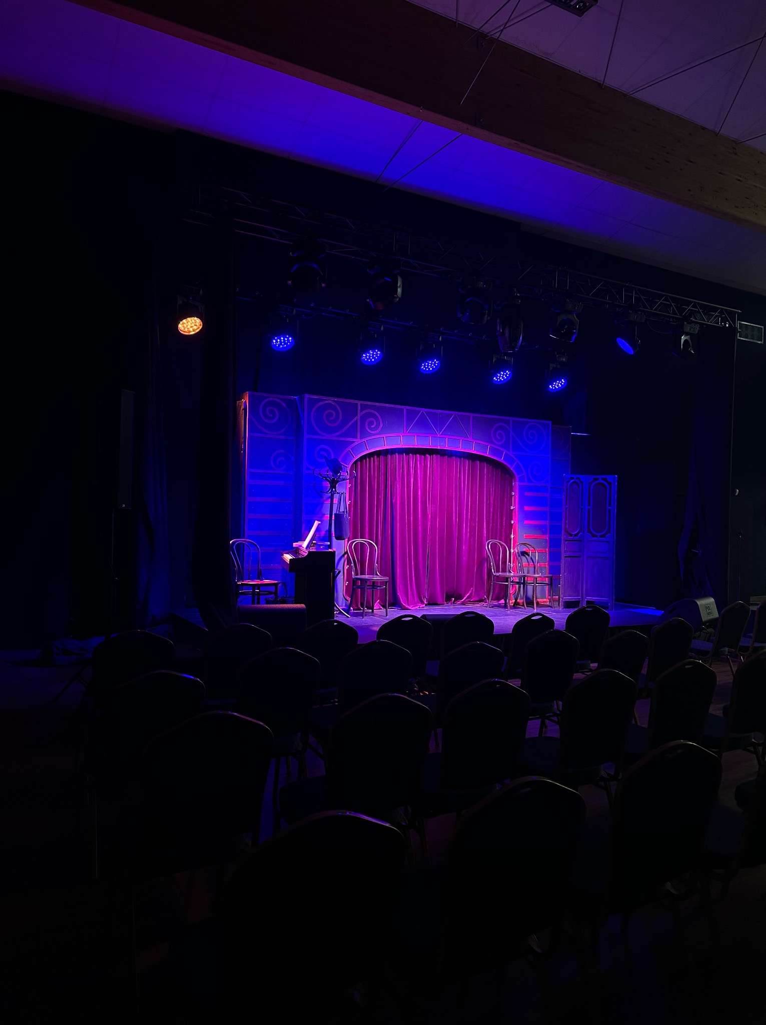 Pusta scena teatralna z fioletowymi zasłonami, krzesłami i pianinem oświetlona reflektorami w kolorze niebieskim i pomarańczowym.