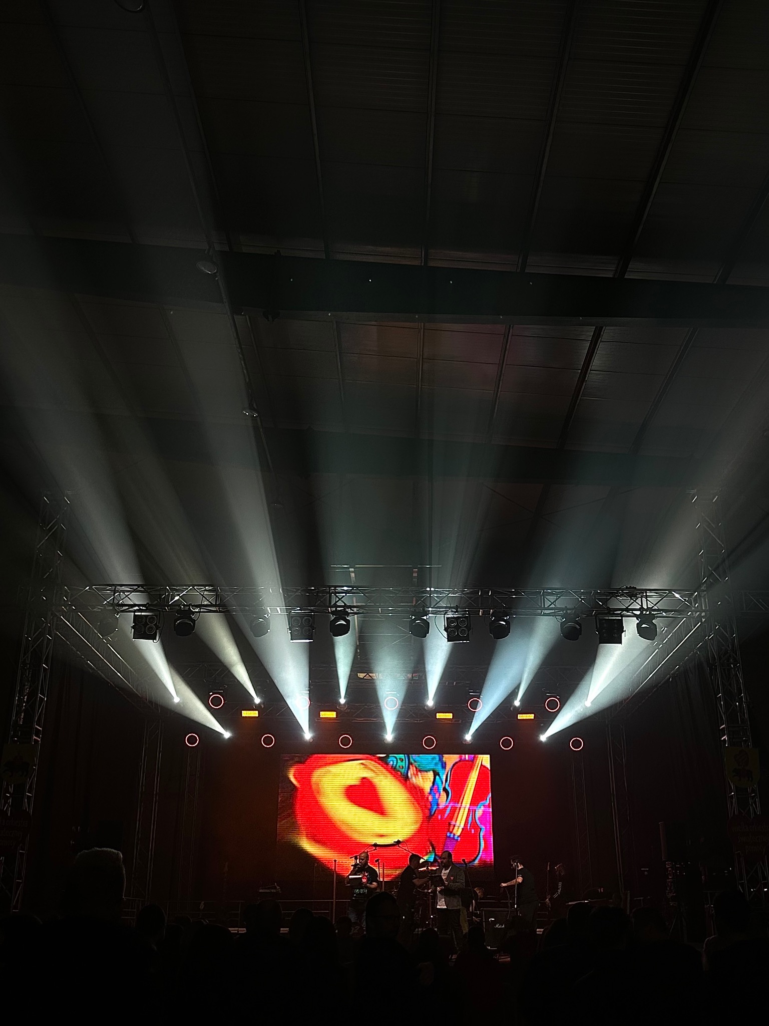 Scena koncertowa z zespołem muzycznym oświetlona reflektorami, z dużym ekranem LED w tle wyświetlającym abstrakcyjną grafikę w odcieniach czerwieni, żółci i błękitu. Reflektory tworzą wyraźne...