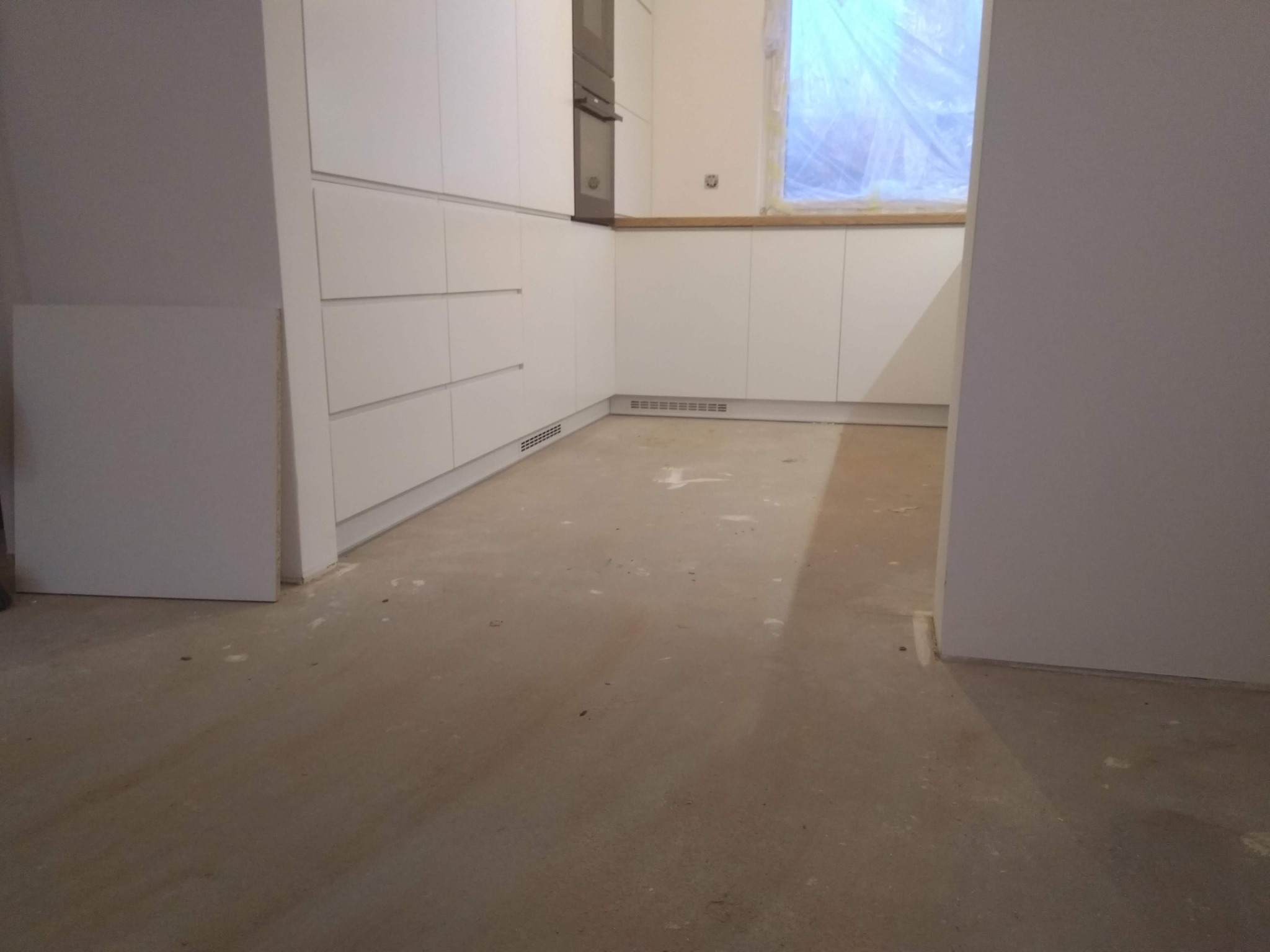 Wnętrze w trakcie wykańczania z zamontowanymi białymi szafkami kuchennymi, piekarnikiem w zabudowie i oknem zabezpieczonym folią malarską.