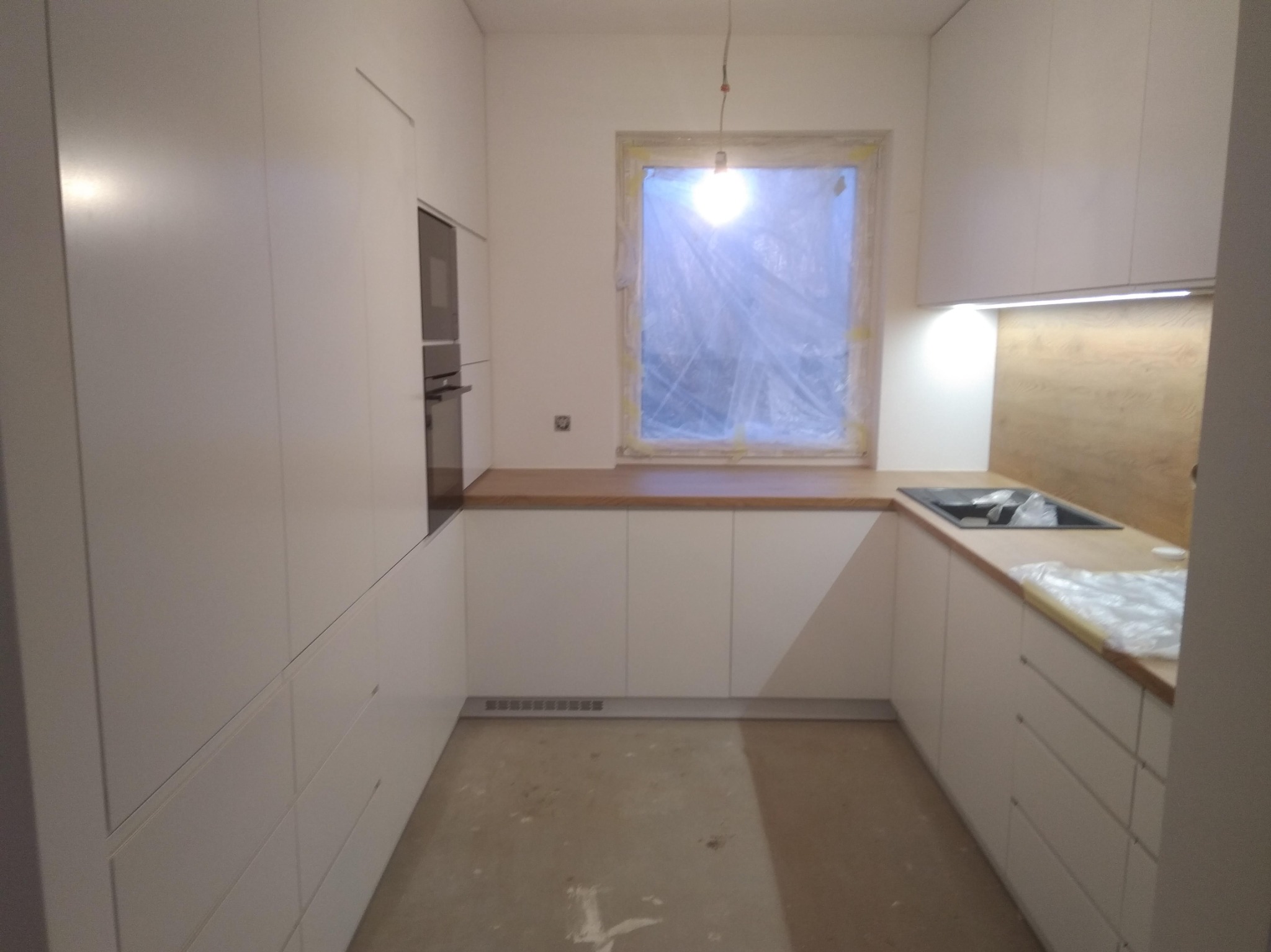 Nowoczesna, biała zabudowa kuchenna z drewnianym blatem w Zalasewie, z oknem zasłoniętym folią ochronną i oświetleniem LED pod szafkami.