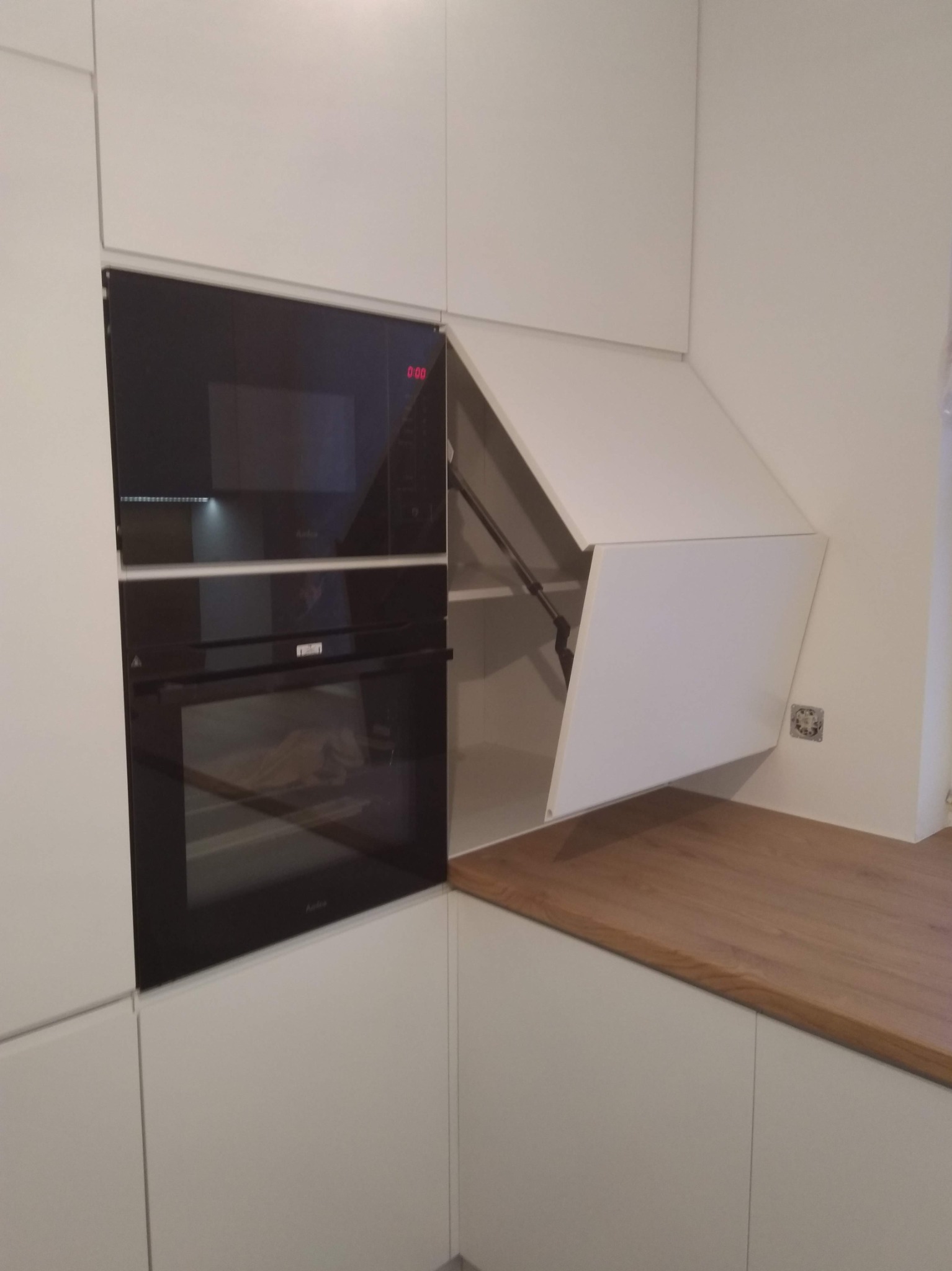 Biała zabudowa kuchenna z piekarnikiem i mikrofalówką w czarnej obudowie; szafka górna z systemem otwierania do góry, blat drewniany.