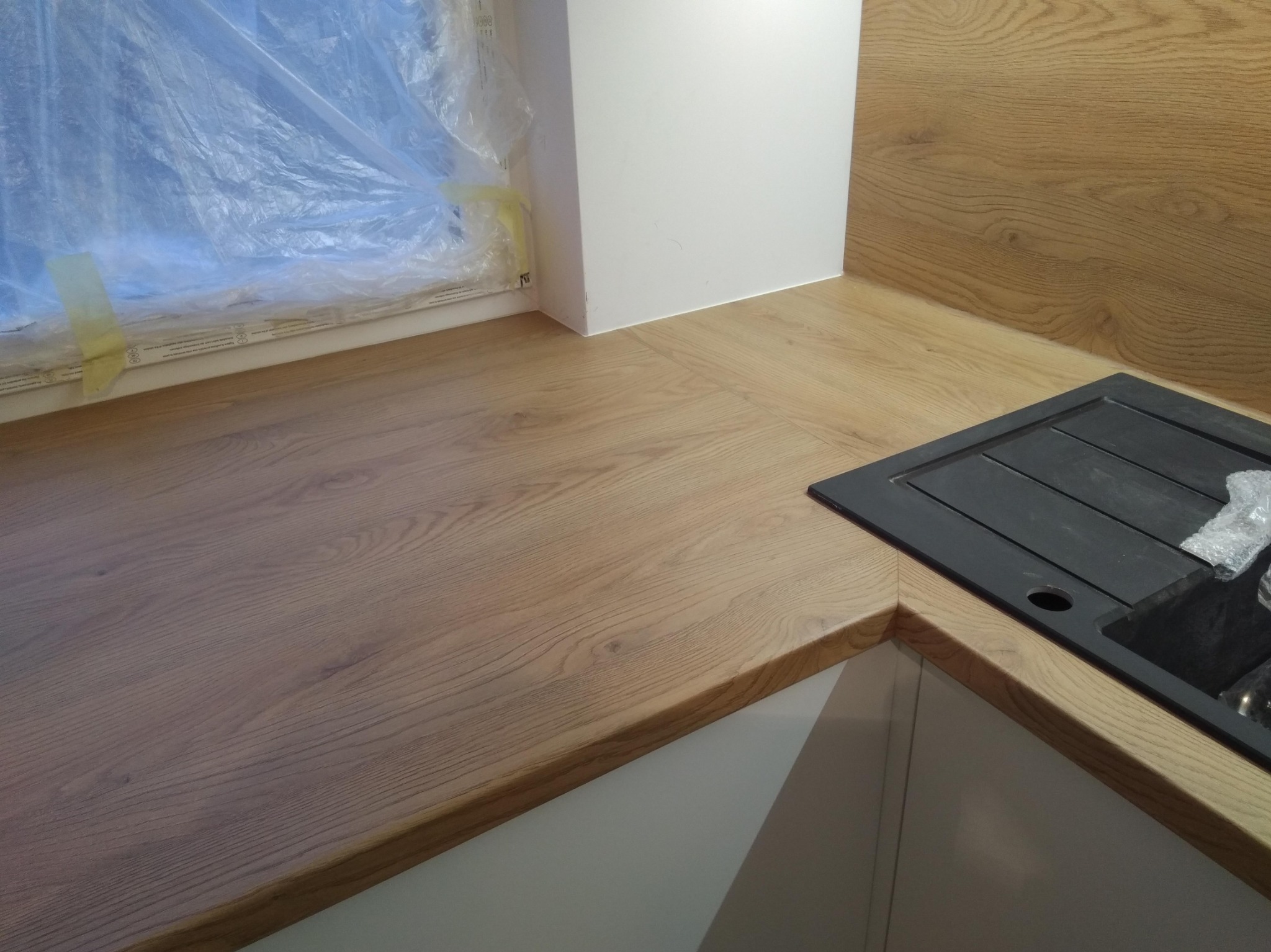 Fragment blatu drewnianego z wbudowanym czarnym zlewozmywakiem, okno zabezpieczone folią malarską, widoczne białe szafki kuchenne.