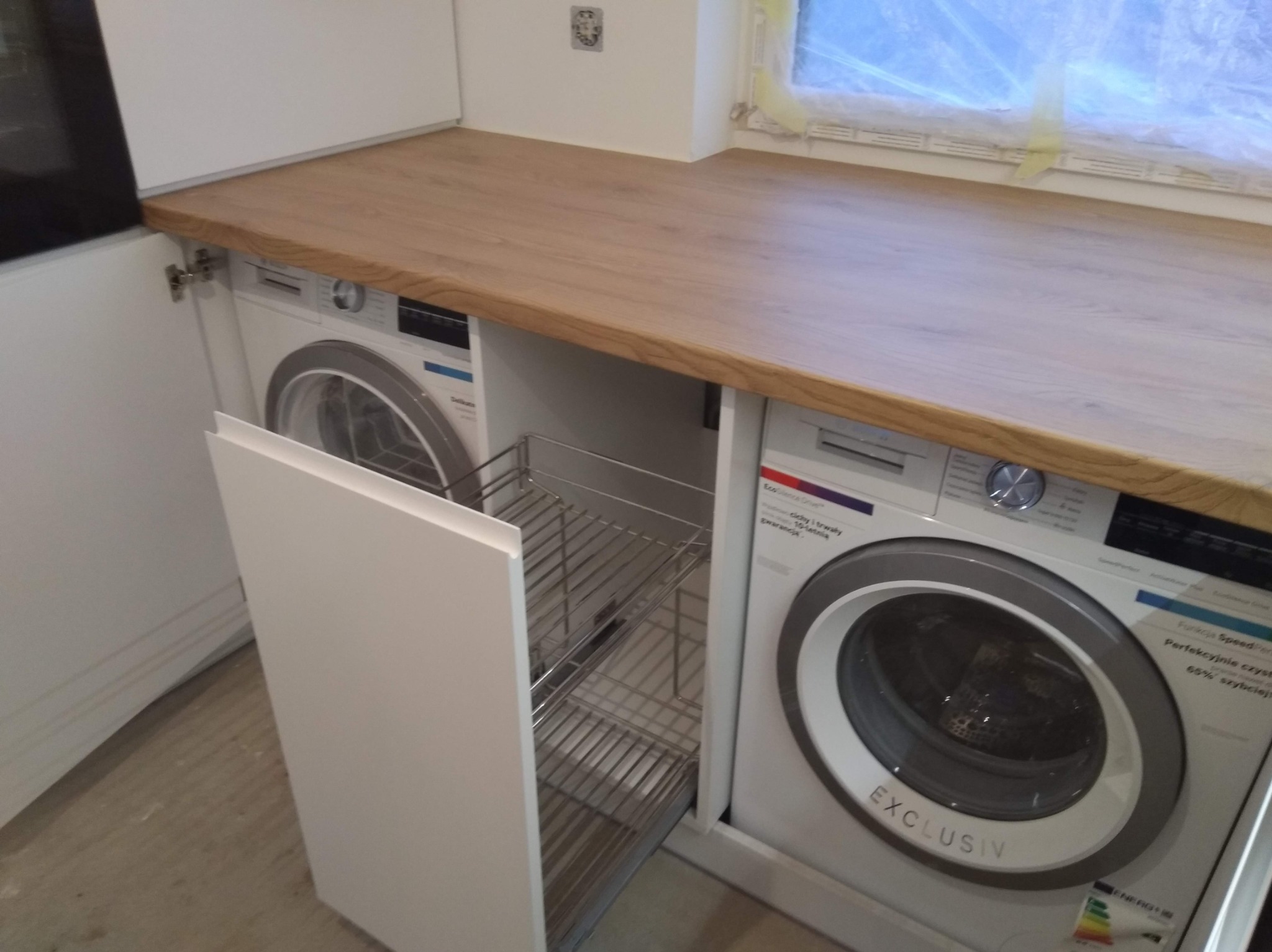 Biała zabudowa kuchenna z drewnianym blatem, integrująca pralki automatyczne z wysuwanym koszem cargo na środki czystości. Okno zasłonięte folią ochronną.