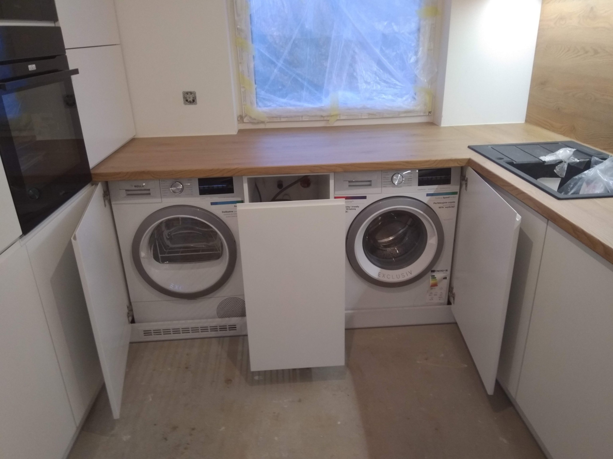 Biała kuchnia na wymiar z drewnianym blatem, zabudowanymi pralką i suszarką za frontami szafek, okno z widocznym zabezpieczeniem remontowym.