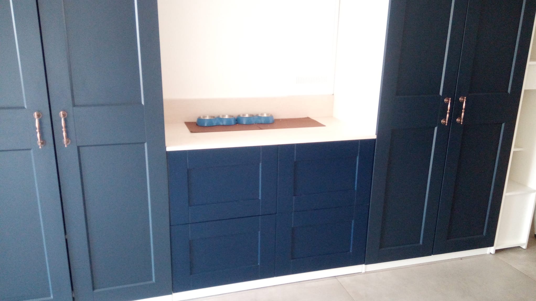 Niebieskie szafki z frezowanymi frontami i miedzianymi uchwytami, część zabudowy z jasnym blatem i miskami dla zwierząt.