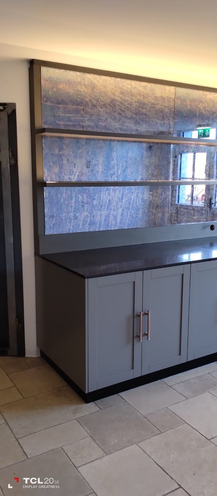 Szara szafka podblatowa z ciemnym blatem i lustrzanymi panelami na górze, wyposażona w miedziane uchwyty, na tle jasnej podłogi z płytek.