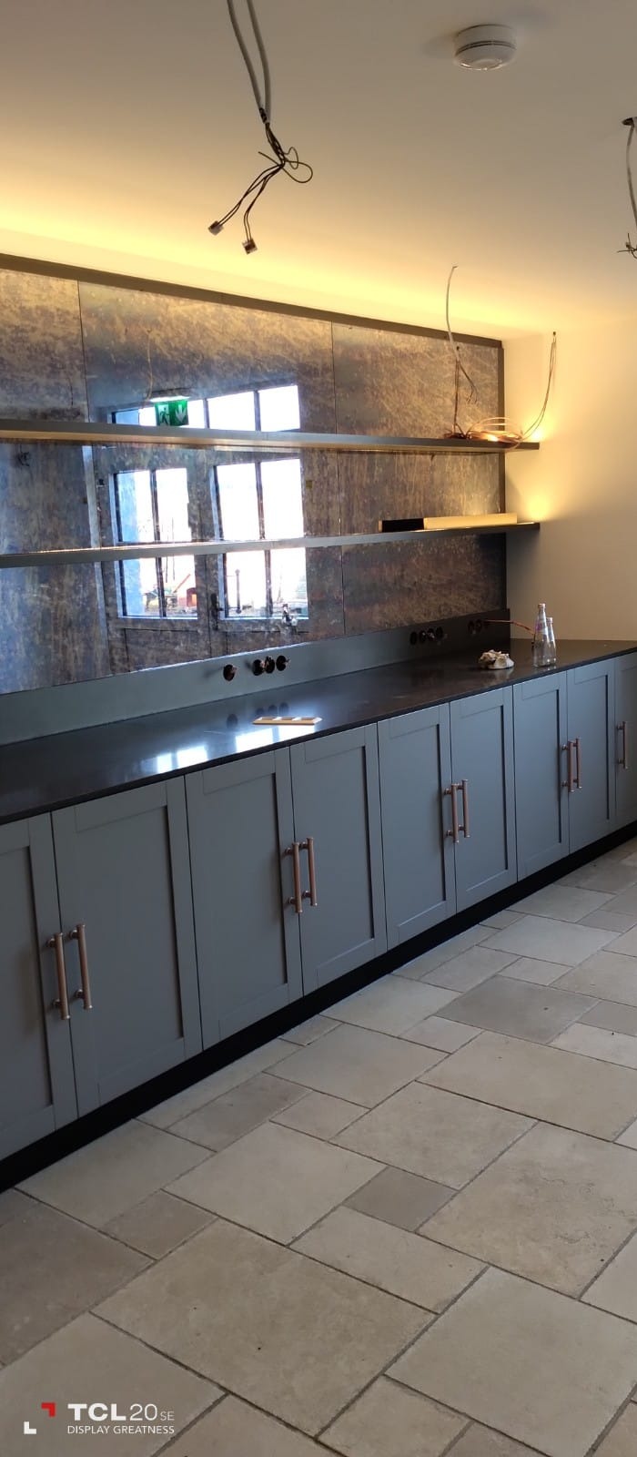 Długa szafka z szarymi frontami i miedzianymi uchwytami pod blatem z ciemnego kamienia. Nad blatem lustrzana ściana z widocznym odbiciem okna i fragmentem sufitu z wystającymi kablami.