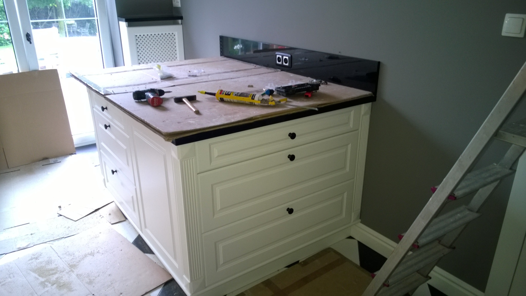 Wnętrze w trakcie montażu wyspy kuchennej w Zalasewie, widoczne narzędzia i materiały montażowe na blacie, białe fronty szuflad w stylu klasycznym, czarny panel ścienny z gniazdkami elektrycznymi.