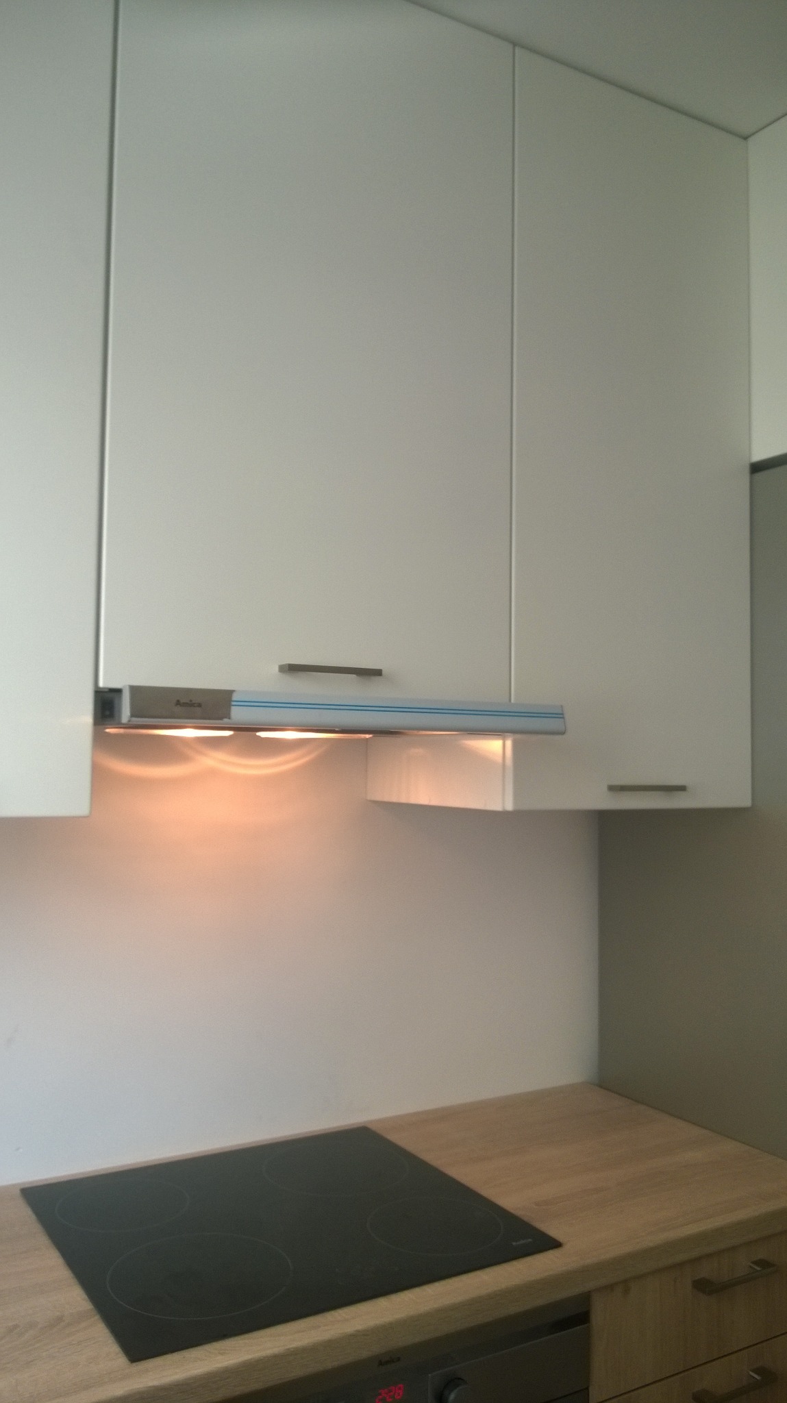 Nowoczesna kuchnia z białymi szafkami wiszącymi, drewnianym blatem i czarną płytą indukcyjną, oświetlona lampkami podszafkowymi.