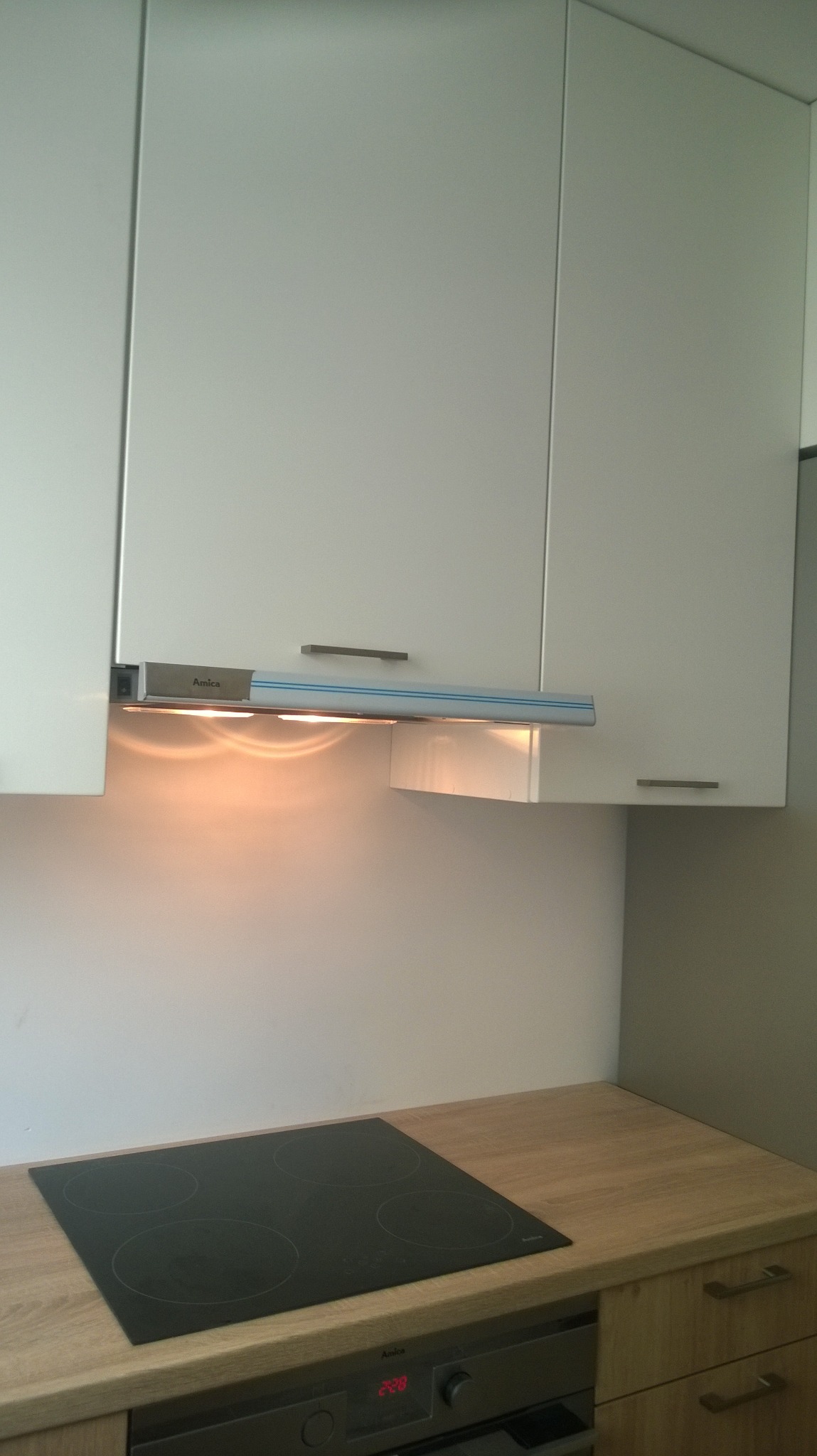 Fragment kuchni z białymi szafkami wiszącymi, okapem z oświetleniem LED marki Amica i płytą indukcyjną na blacie o drewnianej fakturze.