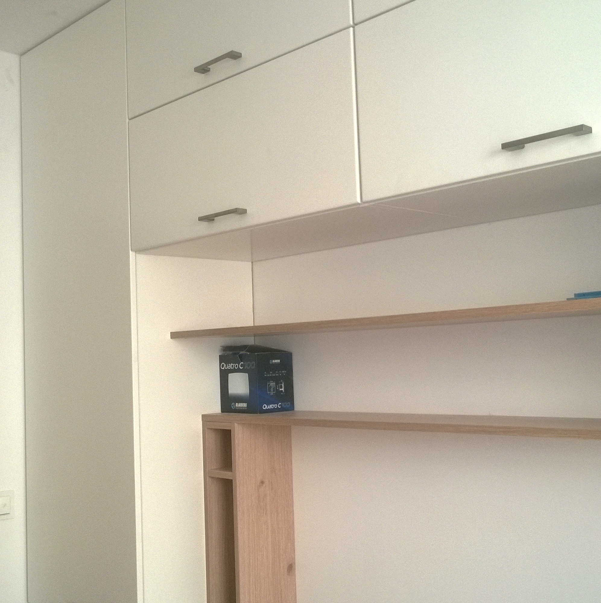 Białe szafki wiszące z metalowymi uchwytami nad drewnianymi półkami, na jednej z nich stoi pudełko.