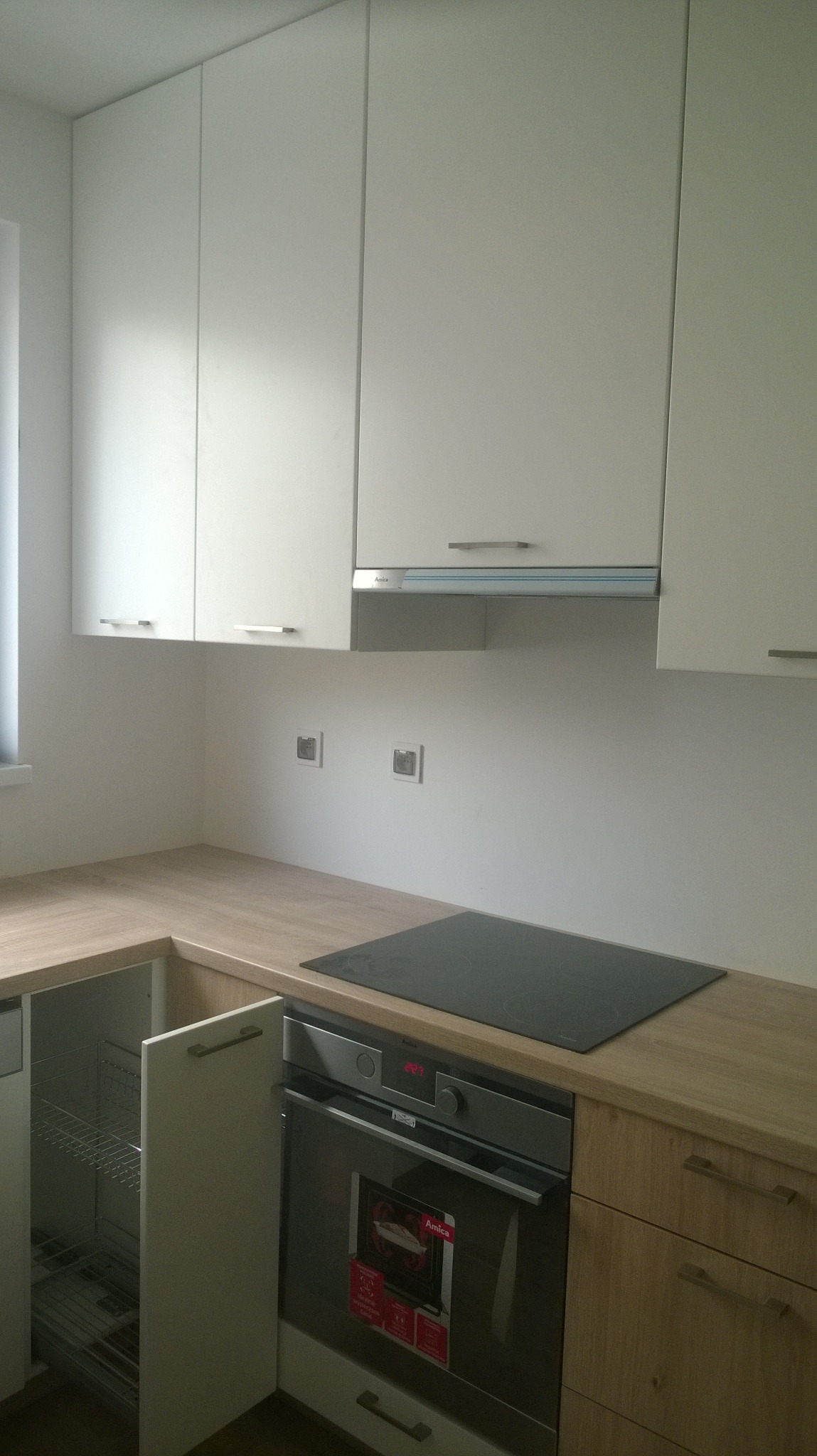 Nowoczesna kuchnia z jasnymi, gładkimi frontami szafek wiszących i drewnianym blatem. Widoczna płyta indukcyjna, piekarnik Amica oraz wysuwana szafka cargo z metalowym koszem.