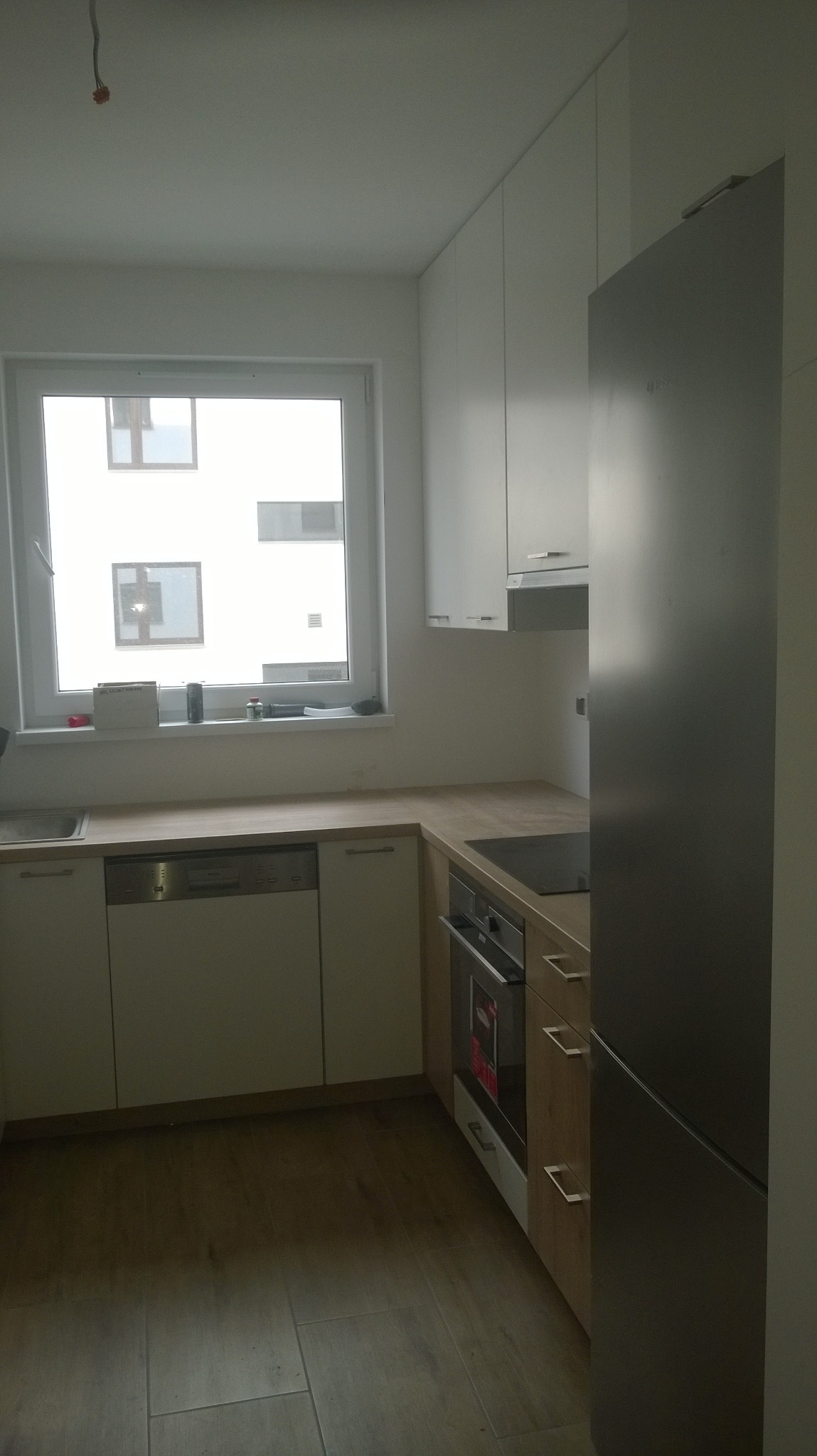 Nowoczesna, jasna kuchnia z białymi szafkami górnymi i dolnymi z drewnianymi akcentami, widok na okno z widokiem na budynek mieszkalny, srebrna lodówka po prawej stronie kadru.