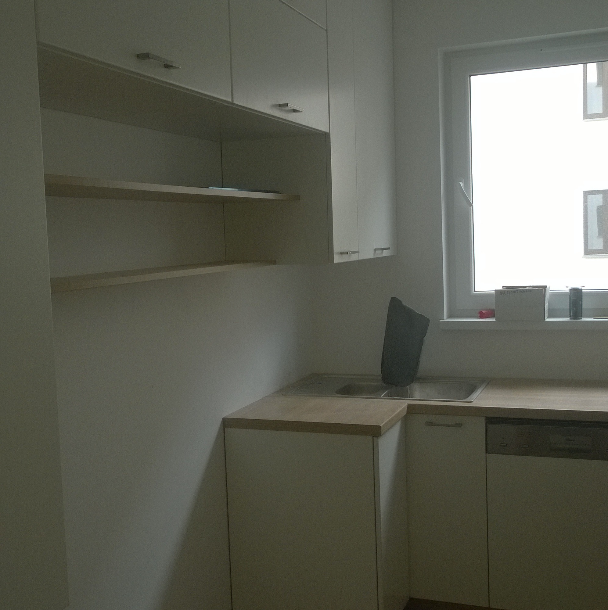 Jasna kuchnia z białymi szafkami, drewnianym blatem i otwartymi półkami, widok na okno z pudełkami i puszką na parapecie.