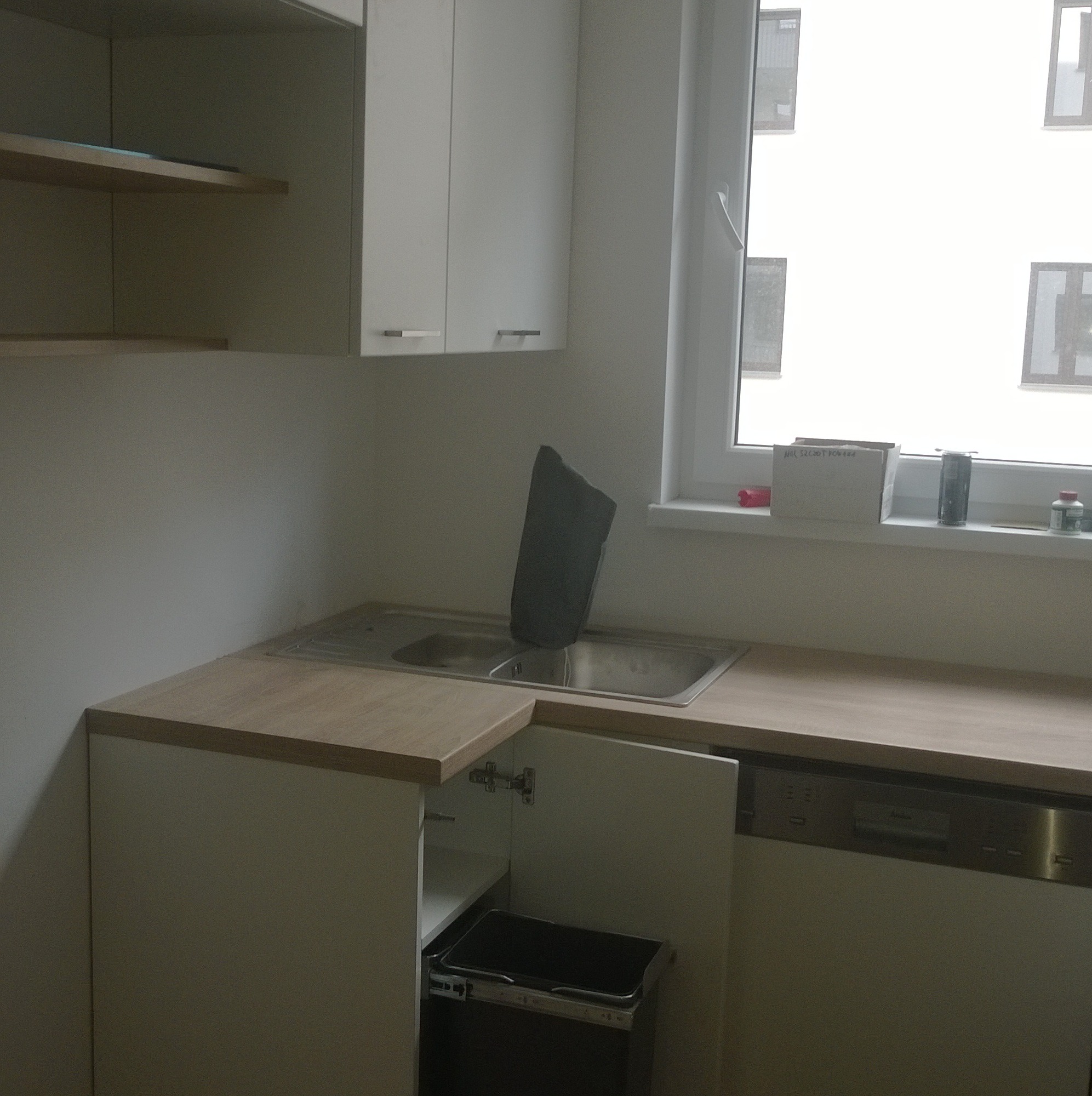 Fragment zabudowy kuchennej z białymi szafkami, drewnianym blatem, zlewozmywakiem z czarną płytą oraz otwartą szafką z wysuwanym koszem na śmieci.