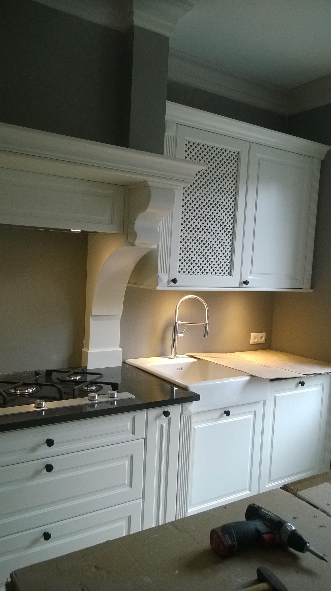 Klasyczna biała kuchnia z czarnym blatem, widoczny zlew typu farmhouse, płyta gazowa, szafka z kratką i narzędzia na kartonie sugerujące montaż.