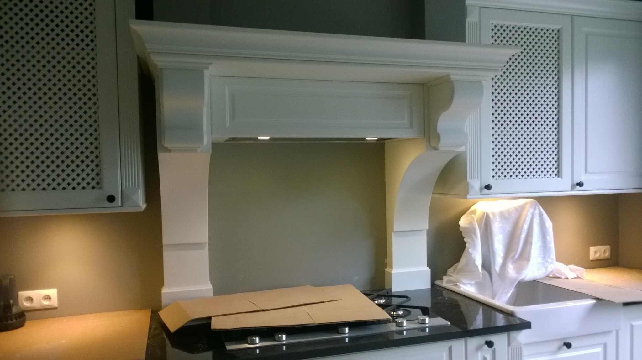 Biała zabudowa kuchenna z ozdobnymi szafkami z kratką, okapem z oświetleniem i czarnym blatem, częściowo zakryta kartonami i tkaniną, widoczne gniazdko elektryczne.