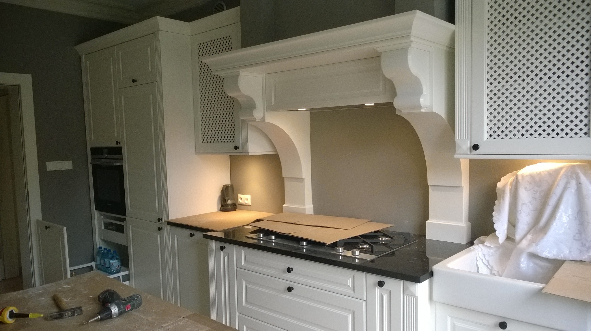 Klasyczna biała kuchnia w trakcie montażu, z widocznymi narzędziami na blacie roboczym, zabezpieczona płyta grzewcza kartonem i przykryty zlew, szafki górne z kratką.
