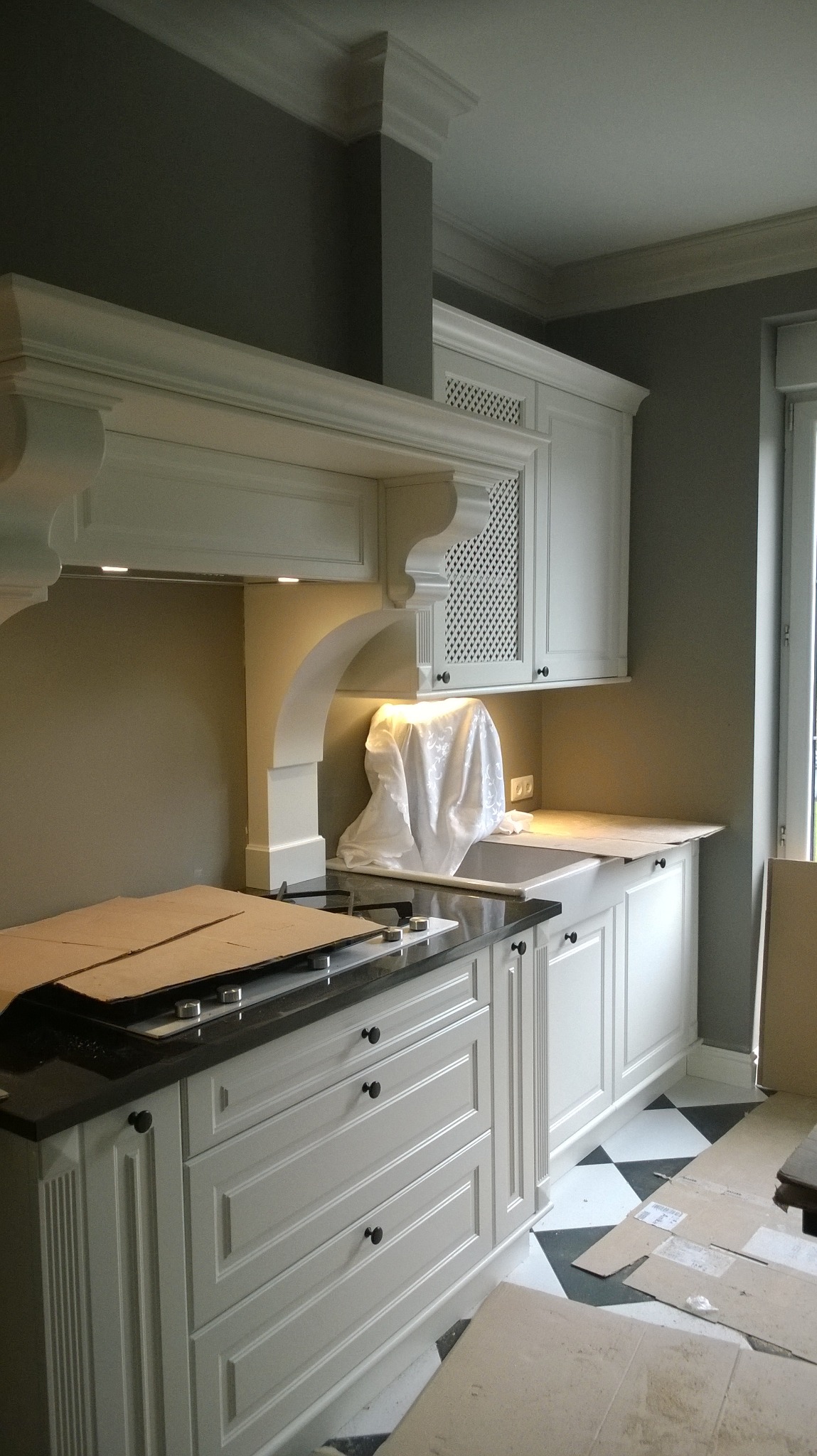 Klasyczna biała zabudowa kuchenna z czarnym blatem, płyta grzewcza zakryta kartonem, zlew przykryty białą tkaniną, podłoga w biało-czarną szachownicę, widoczne elementy wykończeniowe i zabezpieczenia.