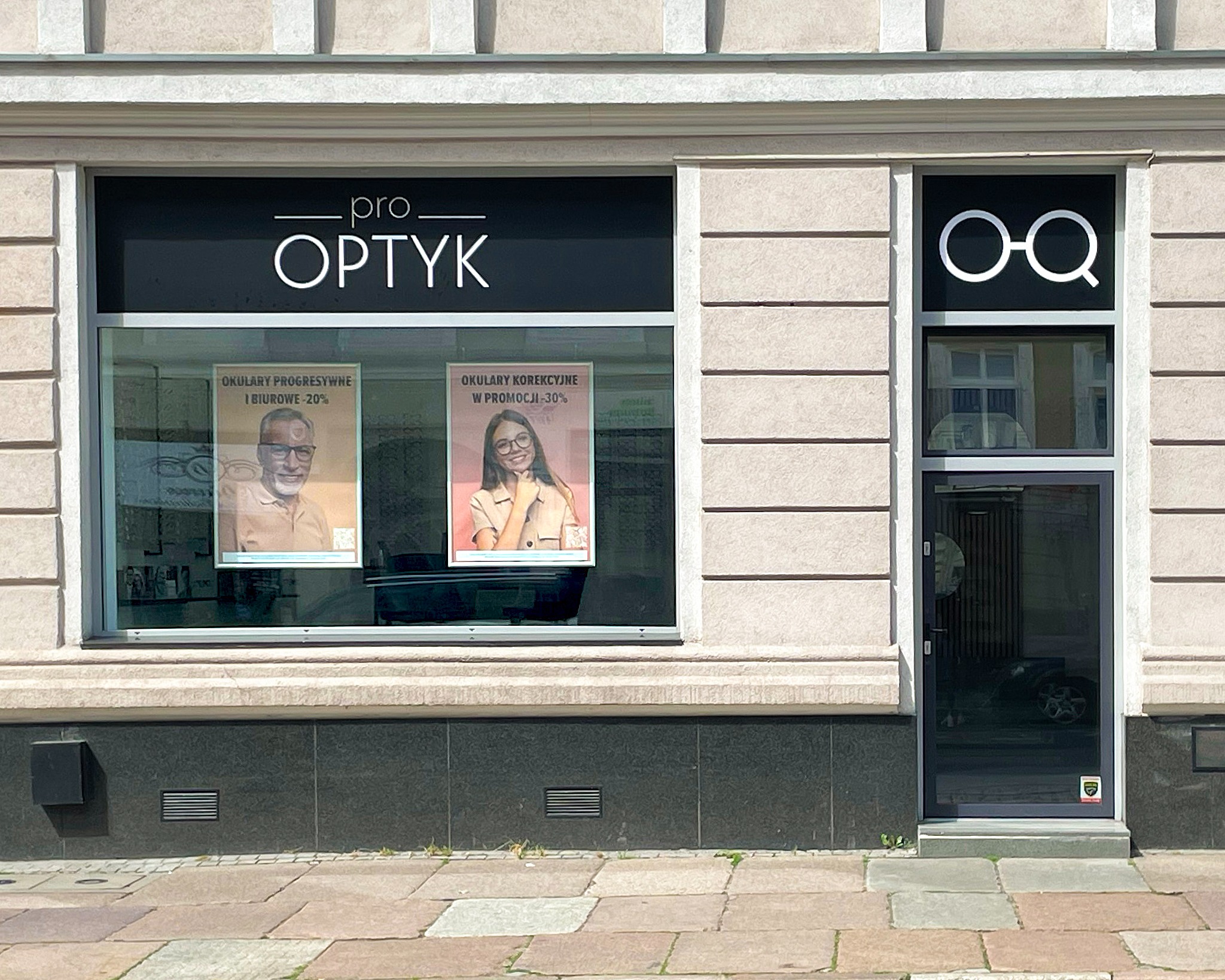 Fasada salonu optycznego z witryną, na której widnieją plakaty reklamowe okularów progresywnych i korekcyjnych w promocji, nad witryną neonowy napis 'pro OPTYK', a obok drzwi z logo okularów...