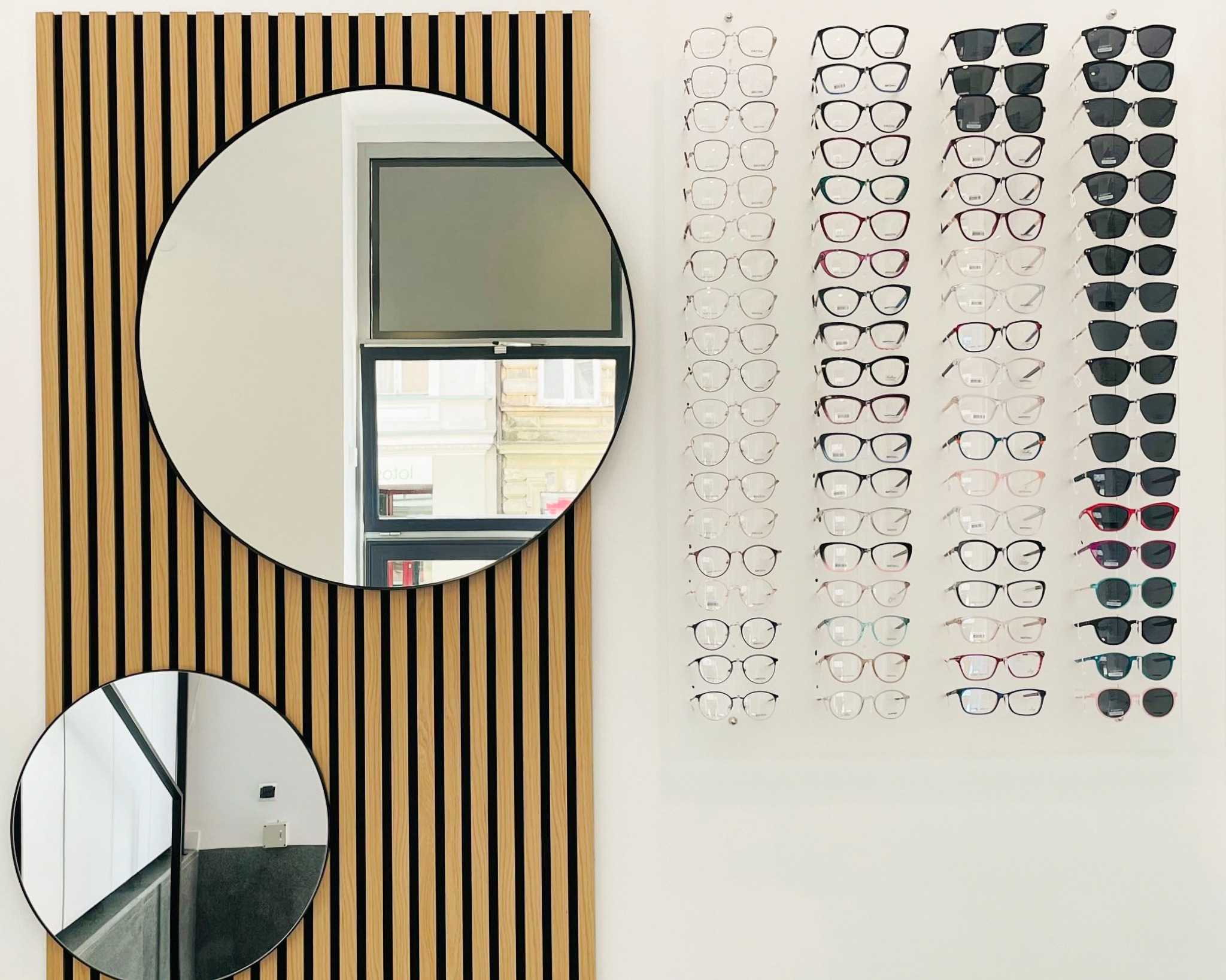 Wystawa oprawek okularowych i okularów przeciwsłonecznych na białej ścianie, obok dekoracyjnej ściany z drewnianych listew i dwóch okrągłych luster.