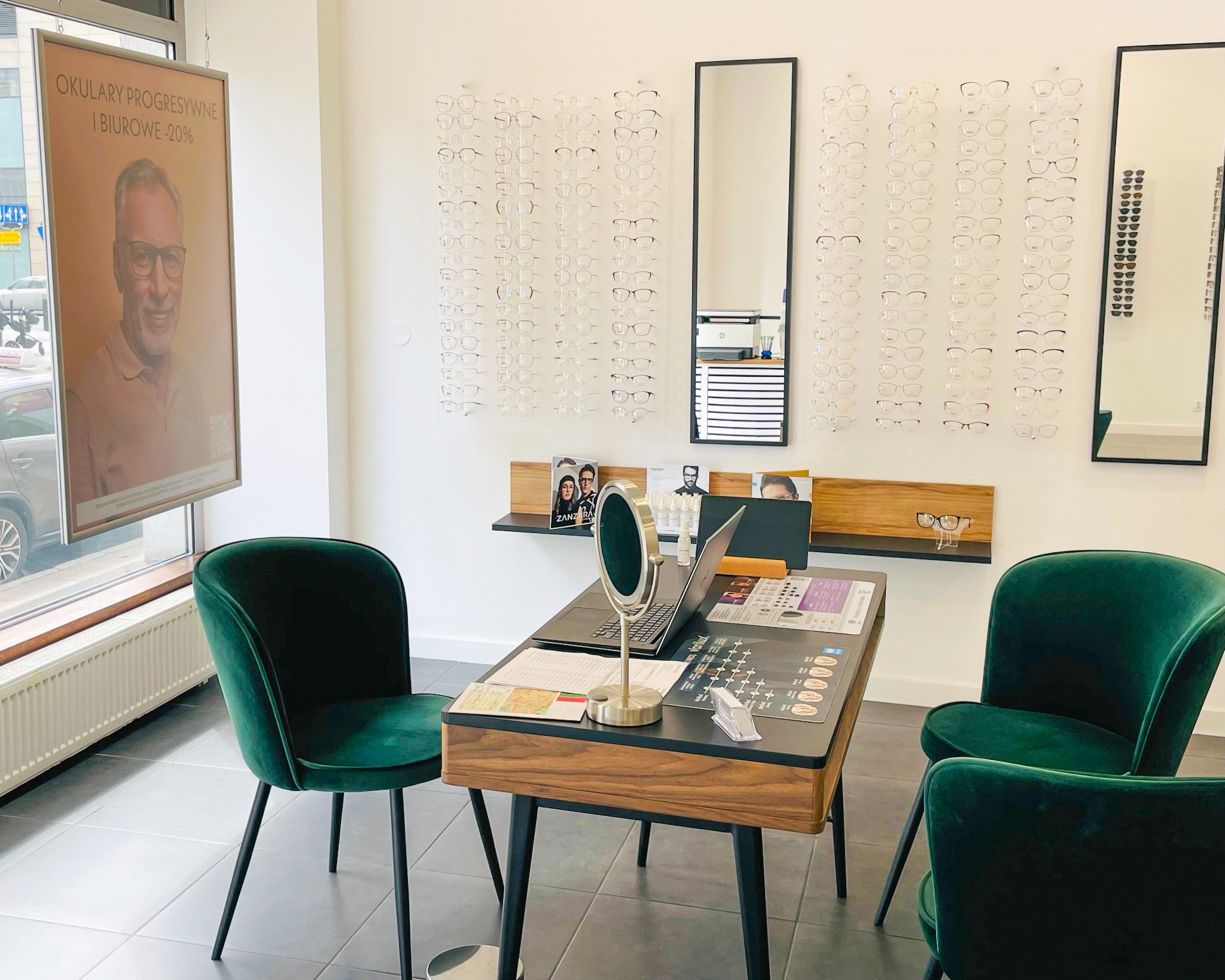 Wnętrze salonu optycznego: ekspozycja okularów na ścianie, stolik z lusterkiem i laptopem, zielone welurowe krzesła, plakat reklamowy okularów progresywnych.