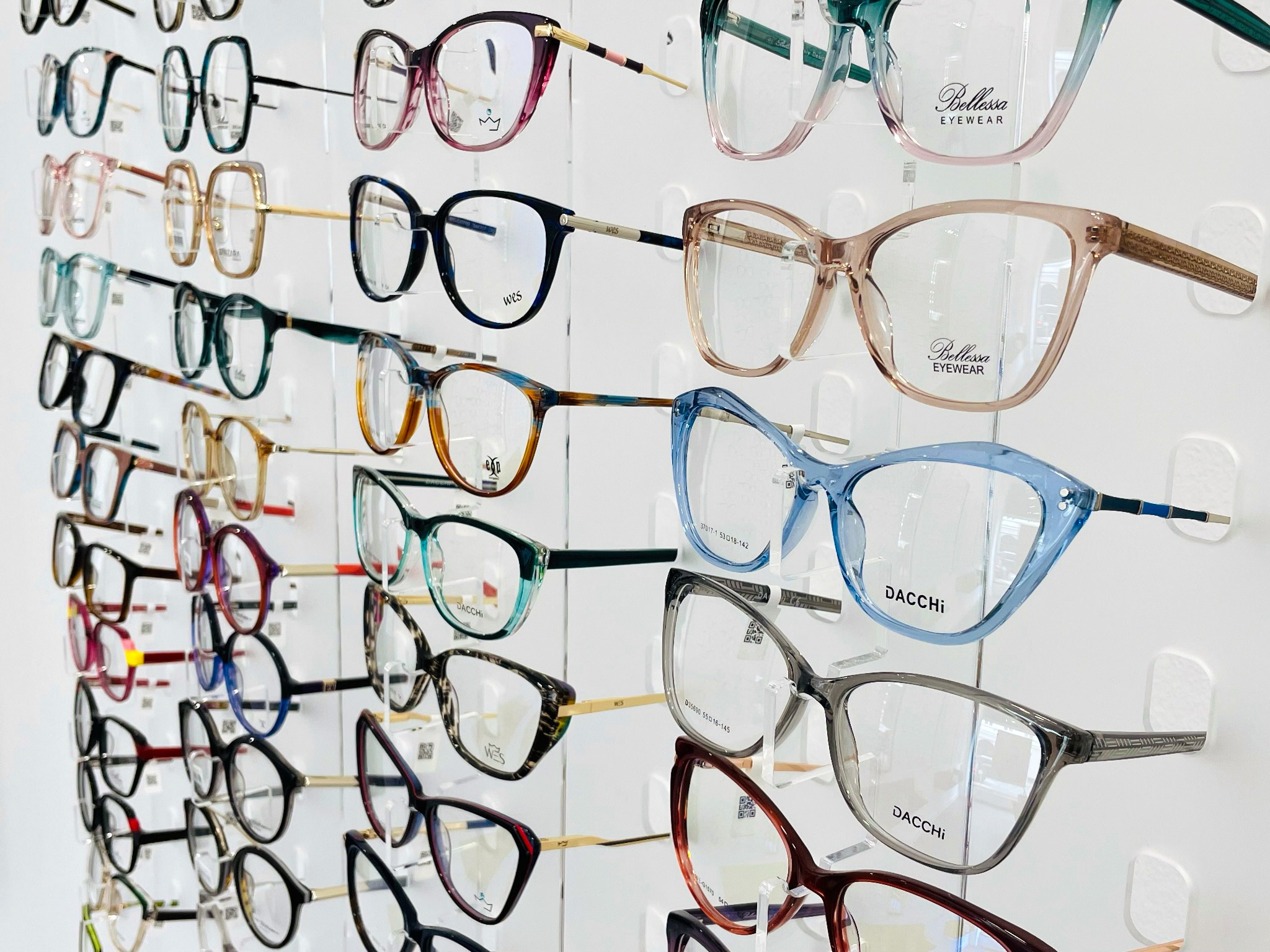 Ekspozycja różnorodnych oprawek okularowych na białej ścianie, modele marek Bellessa i Dacchi, widoczne detale wykończenia zauszników i oznaczenia na szkłach.