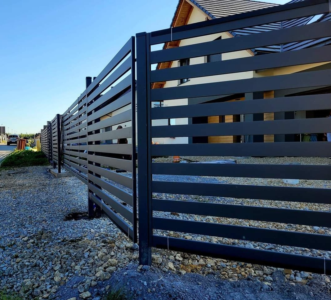 Nowoczesne, poziome, szare ogrodzenie z paneli stalowych przed domem z dwuspadowym dachem i jasną elewacją, widok z poziomu gruntu na żwirowym podłożu, błękitne niebo w tle.