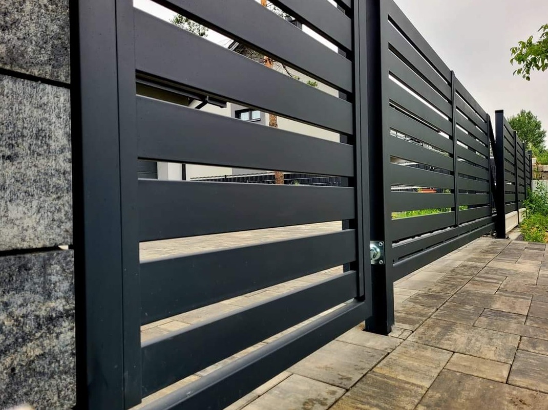 Nowoczesna, przesuwna brama wjazdowa z poziomych, matowych profili w kolorze grafitowym, zamontowana na posesji z brukowanym podjazdem i kamiennym ogrodzeniem.