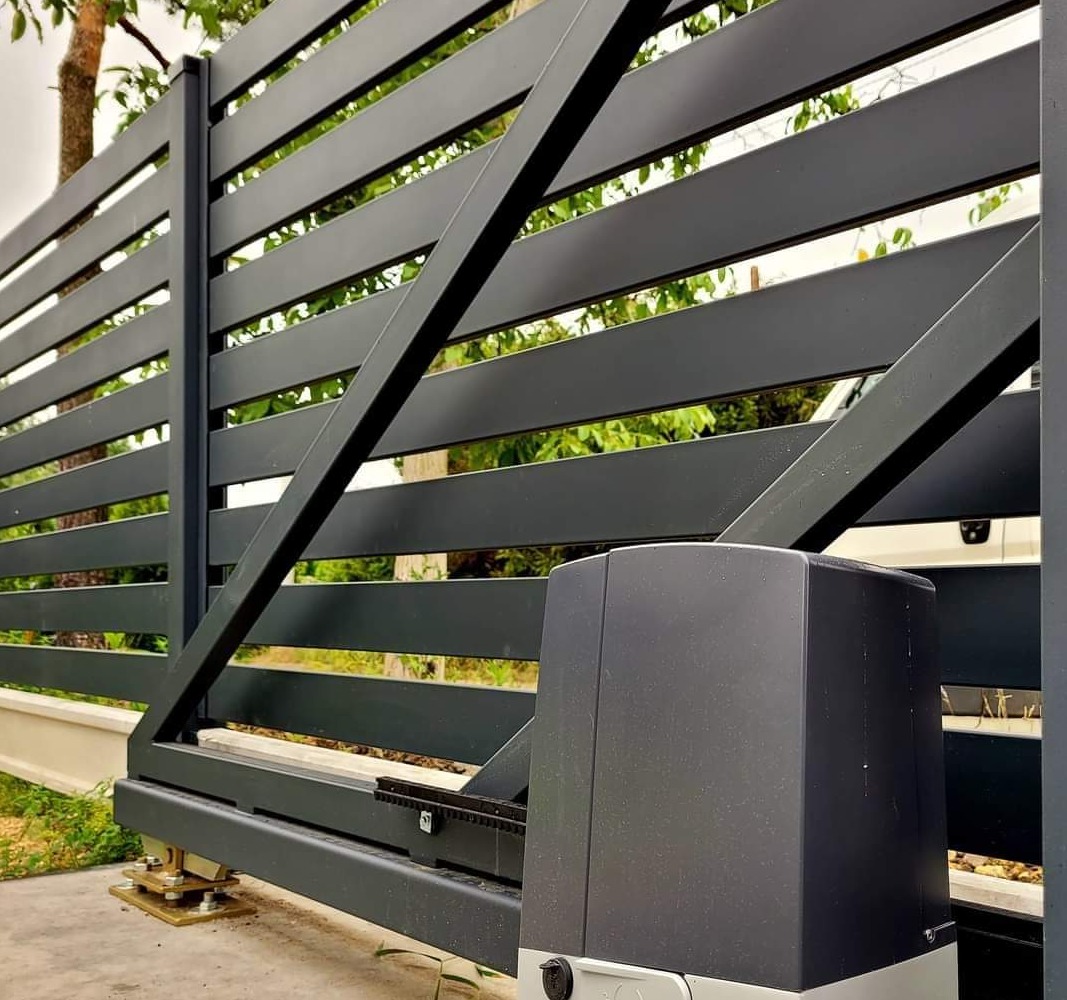 Nowoczesna, przesuwna brama wjazdowa z automatycznym napędem, pomalowana na grafitowy kolor, widoczna na tle zieleni ogrodu i fragmentu białego samochodu.