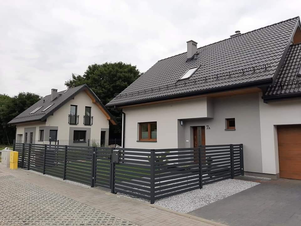 Nowoczesne, poziome ogrodzenie panelowe w kolorze grafitowym przed dwoma domami z szarą elewacją i ciemnymi dachami, widok z poziomu chodnika.