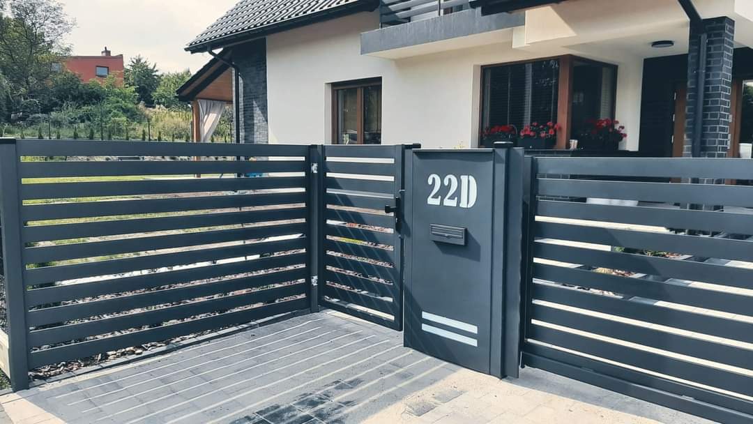 Nowoczesna, pozioma, szara brama wejściowa z numerem 22D i skrzynką na listy, wkomponowana w ogrodzenie z poziomych paneli, na tle domu z białymi ścianami i ciemną dachówką.
