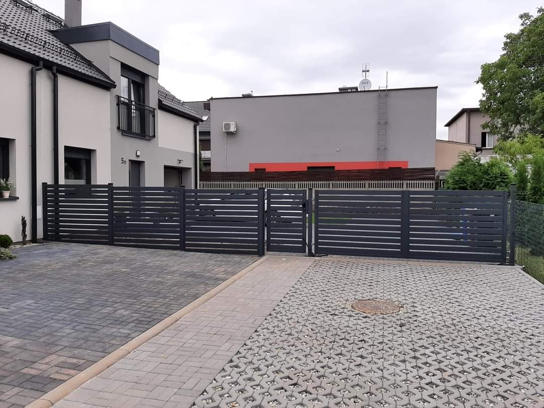 Nowoczesna, pozioma, grafitowa brama wjazdowa i przęsła ogrodzeniowe z poprzecznymi listwami, dopasowane do architektury domu z szarą elewacją i brukowanym podjazdem.