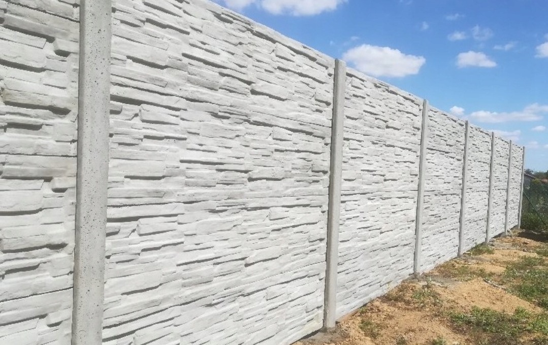 Długi, szary mur z prefabrykowanych paneli betonowych o fakturze imitującej kamień, wsparty na pionowych słupkach, widoczny z boku na tle błękitnego nieba i zieleni.