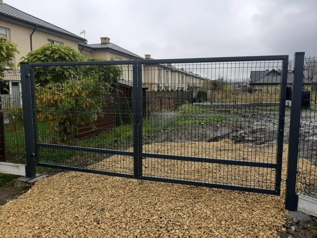 Nowoczesna, dwuskrzydłowa brama z paneli zgrzewanych w kolorze grafitowym, zamontowana na posesji z wysypanym żwirem, widok z zewnątrz.