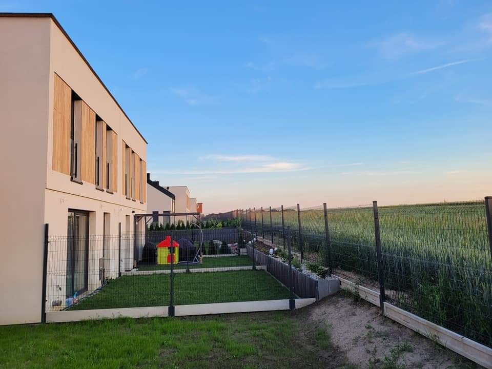 Nowoczesny dom z jasną elewacją i drewnianymi akcentami, otoczony zielonym trawnikiem i niskim ogrodzeniem panelowym w kolorze antracytowym, za którym rozciąga się pole zboża pod błękitnym niebem...