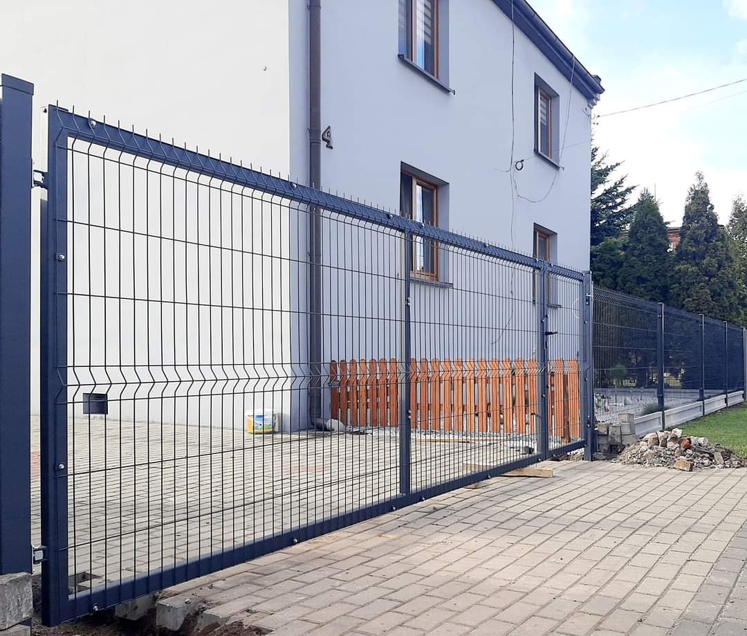Grafitowa brama wjazdowa z paneli ogrodzeniowych na posesji z kostki brukowej, widoczny fragment ogrodzenia panelowego i budynek w tle.