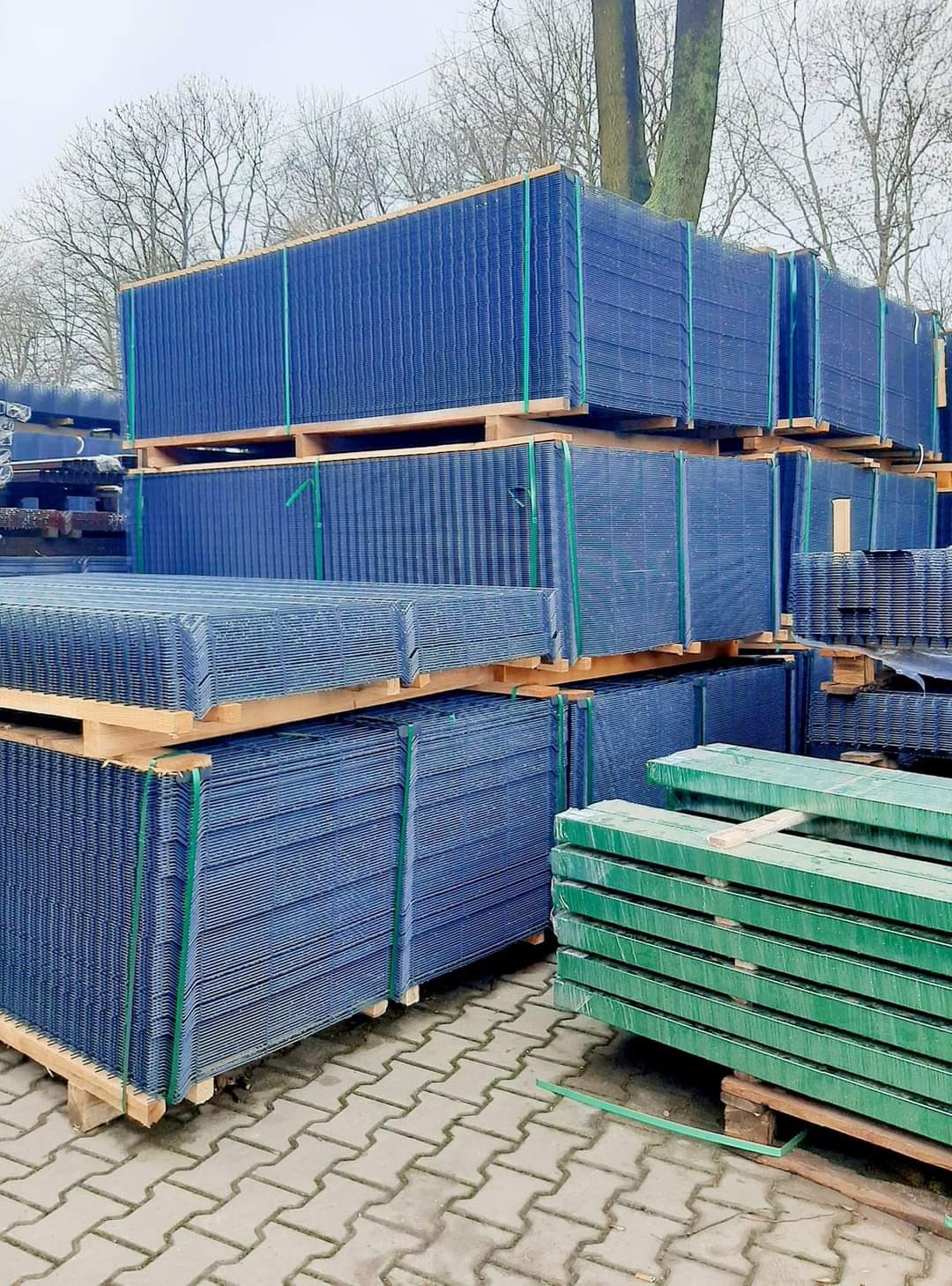 Stosy niebieskich paneli ogrodzeniowych i zielonych słupków na paletach, zabezpieczone taśmami, ułożone na kostce brukowej, widoczne drzewa w tle.