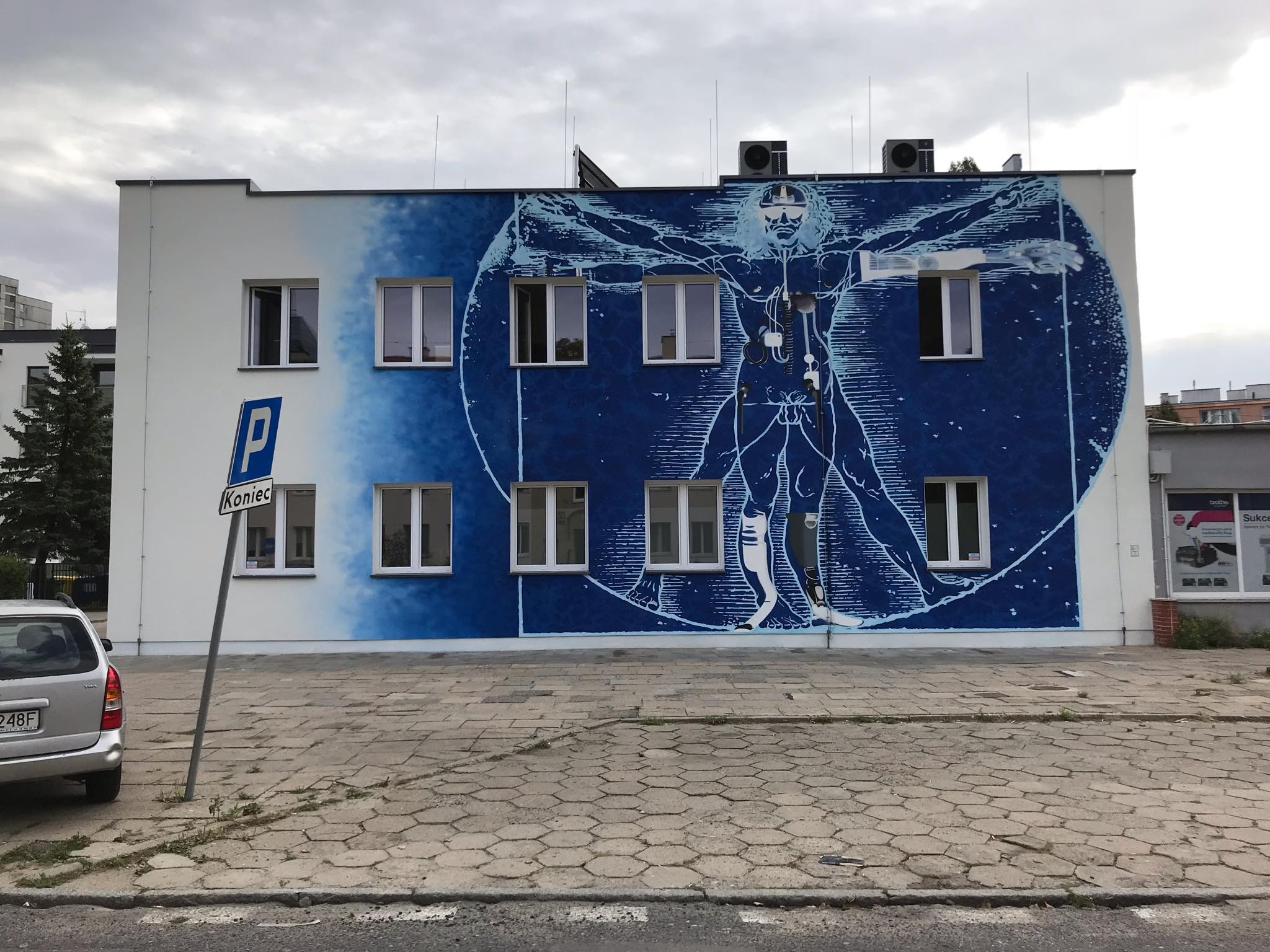 Budynek z muralem przedstawiającym Człowieka Witruwiańskiego w odcieniach błękitu, z oknami wkomponowanymi w projekt, widoczny znak drogowy 'Koniec' i zaparkowany samochód.