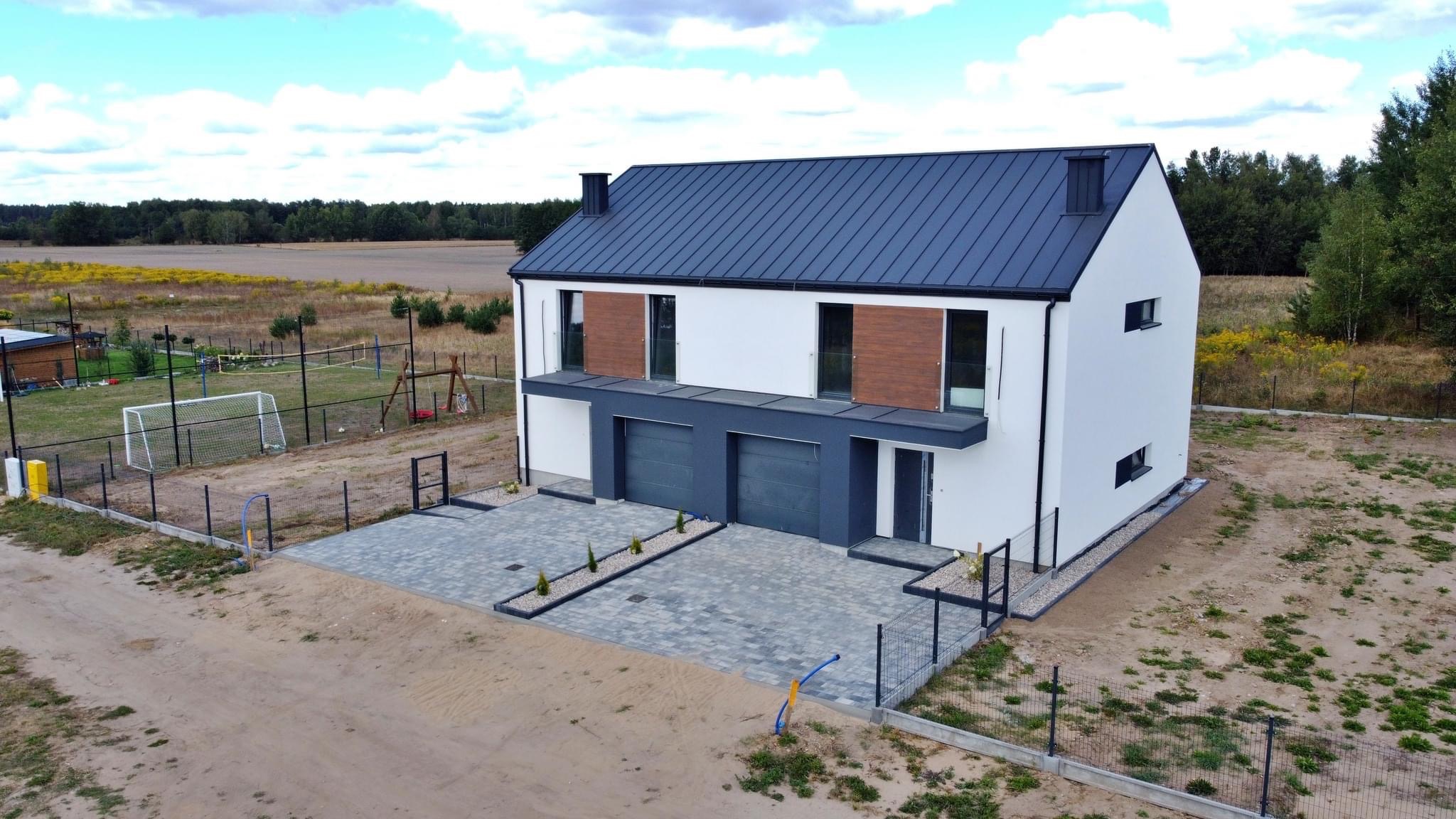 Nowoczesny dwukondygnacyjny dom bliźniak z płaskim dachem, grafitowymi bramami garażowymi i drewnianymi akcentami na fasadzie, widok z lotu ptaka na częściowo zagospodarowaną działkę z widocznym...