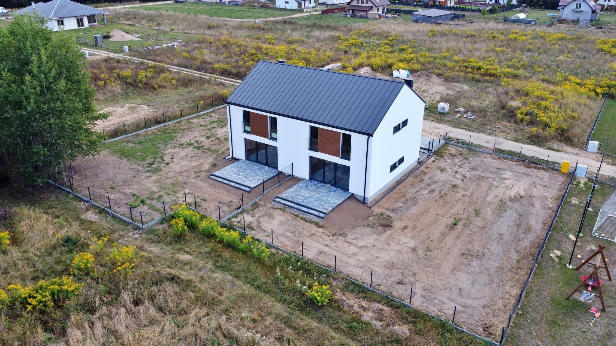 Nowoczesny, dwukondygnacyjny dom bliźniak z białymi ścianami i grafitowym dachem, widziany z lotu ptaka, z przygotowanym terenem wokół i ogrodzeniem z siatki.