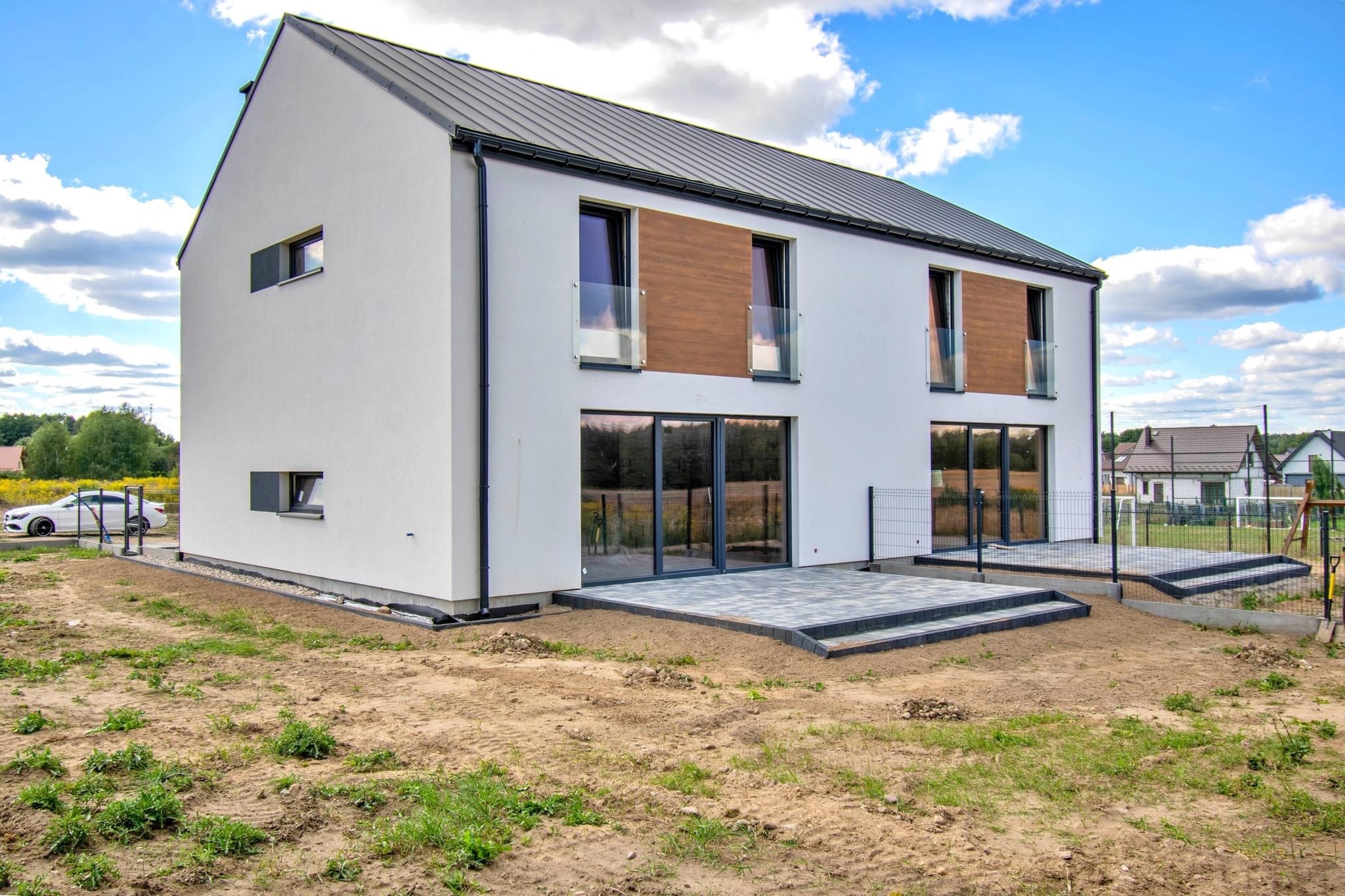 Nowoczesny, dwukondygnacyjny dom z płaskim dachem i białymi ścianami, z drewnianymi akcentami na elewacji, szarymi ramami okiennymi i szklanymi balustradami, świeżo ułożony taras z kostki brukowej...