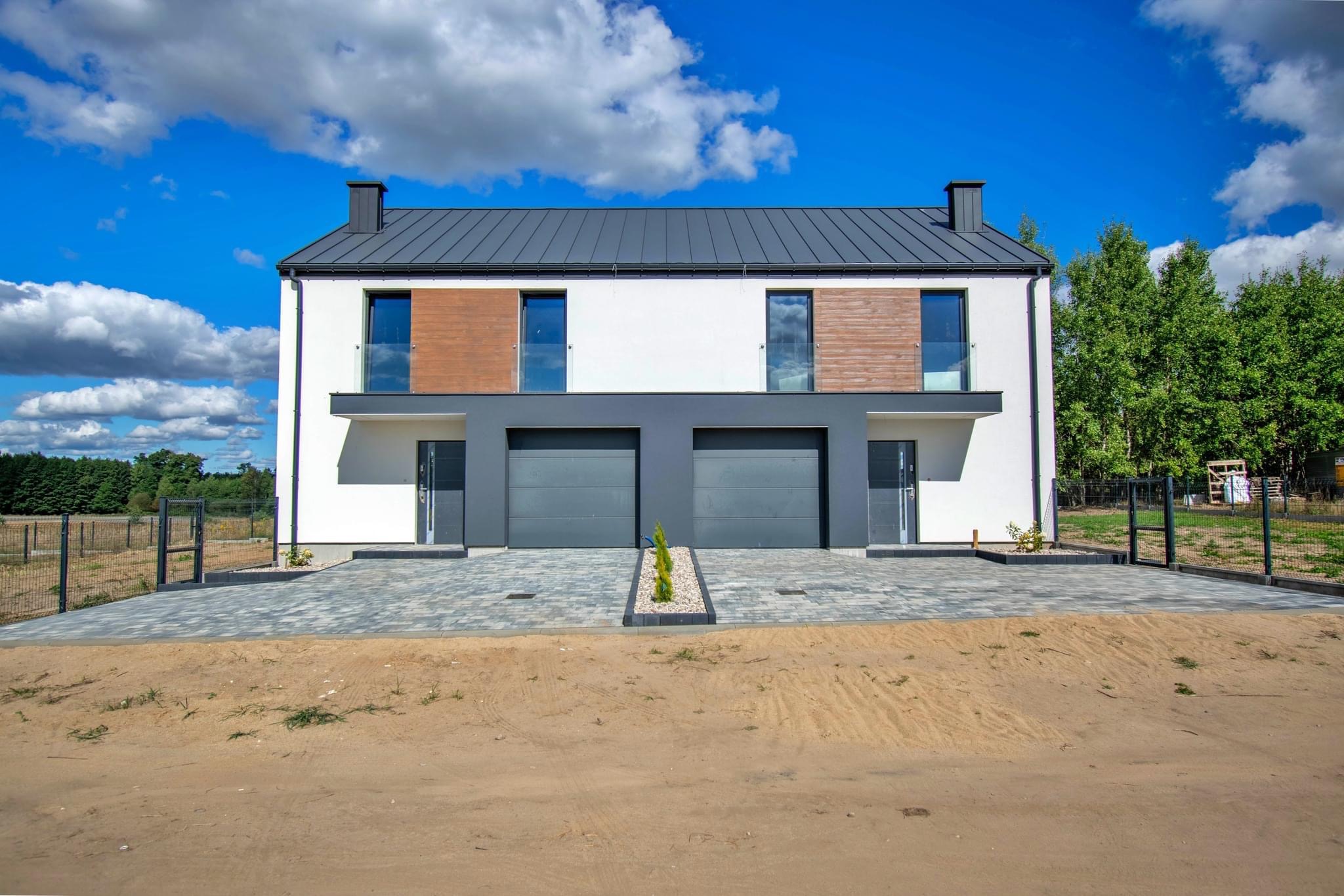 Nowoczesny bliźniak z szarą blachodachówką, drewnianymi panelami elewacyjnymi, dwoma garażami i kostką brukową na podjeździe w słoneczny dzień.