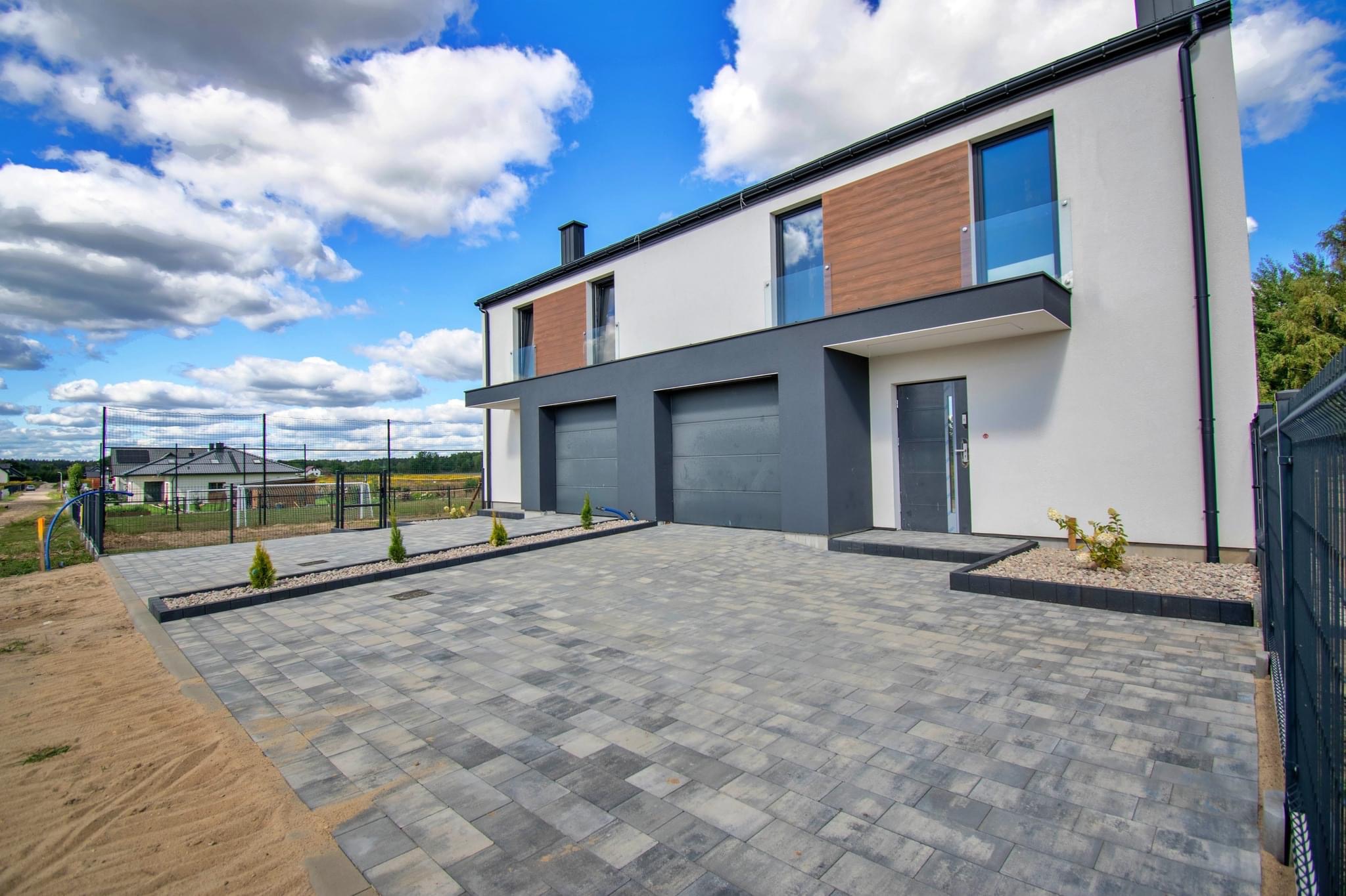 Nowoczesny, dwukondygnacyjny dom z płaskim dachem, szarą elewacją nad garażami i drewnianymi panelami na piętrze, widok z poziomu brukowanego podjazdu z młodymi drzewkami.